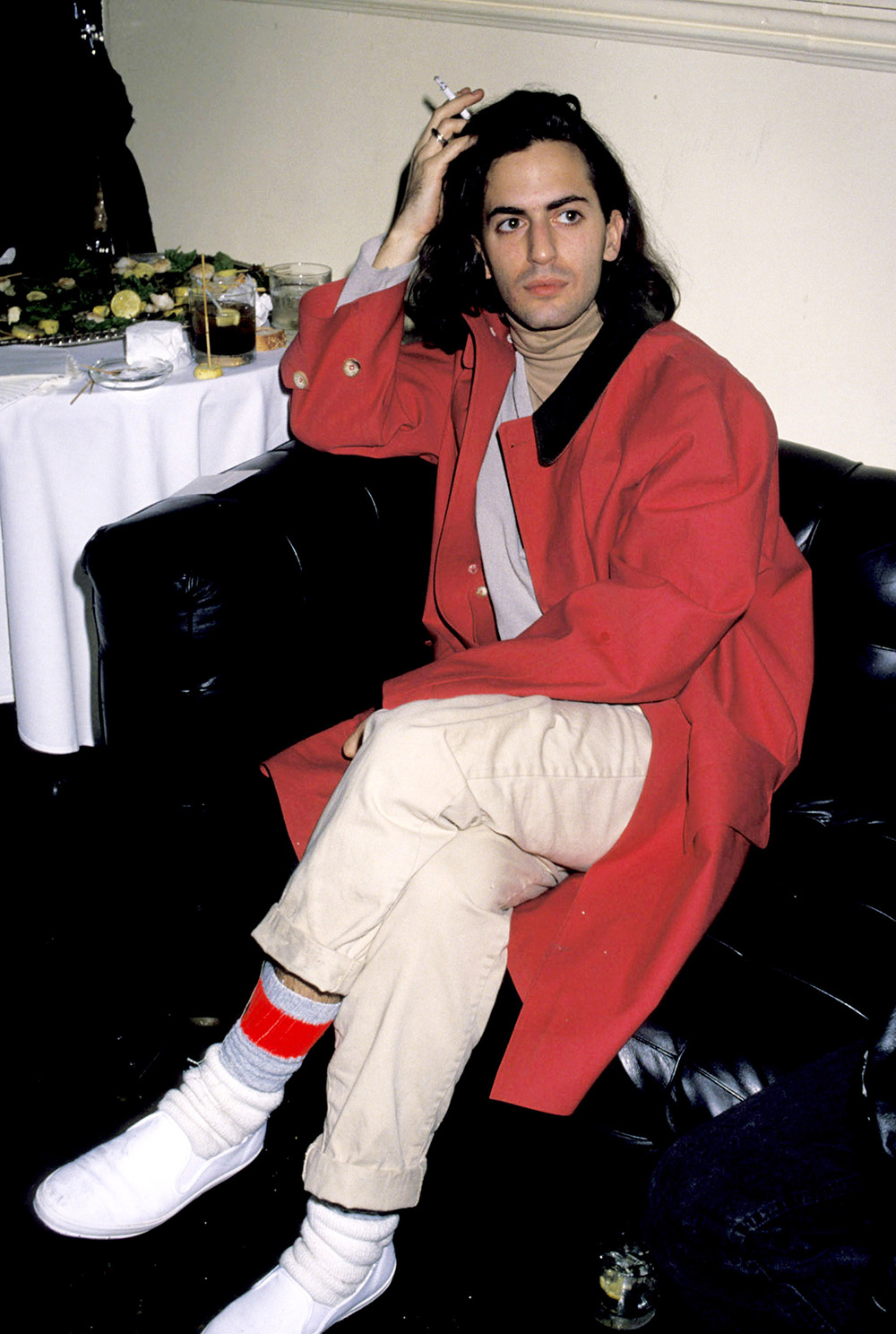 Marc Jacobs w czerwonym płaszczu, siedzi na czarnej kanapie, zapalając papierosa. W tle stół z jedzeniem i napojami.