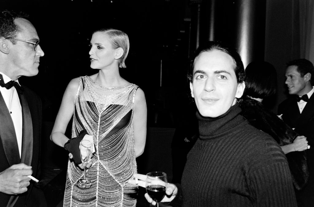 Marc Jacobs stoi z drinkiem w towarzystwie elegancko ubranych gości na wydarzeniu towarzyskim.