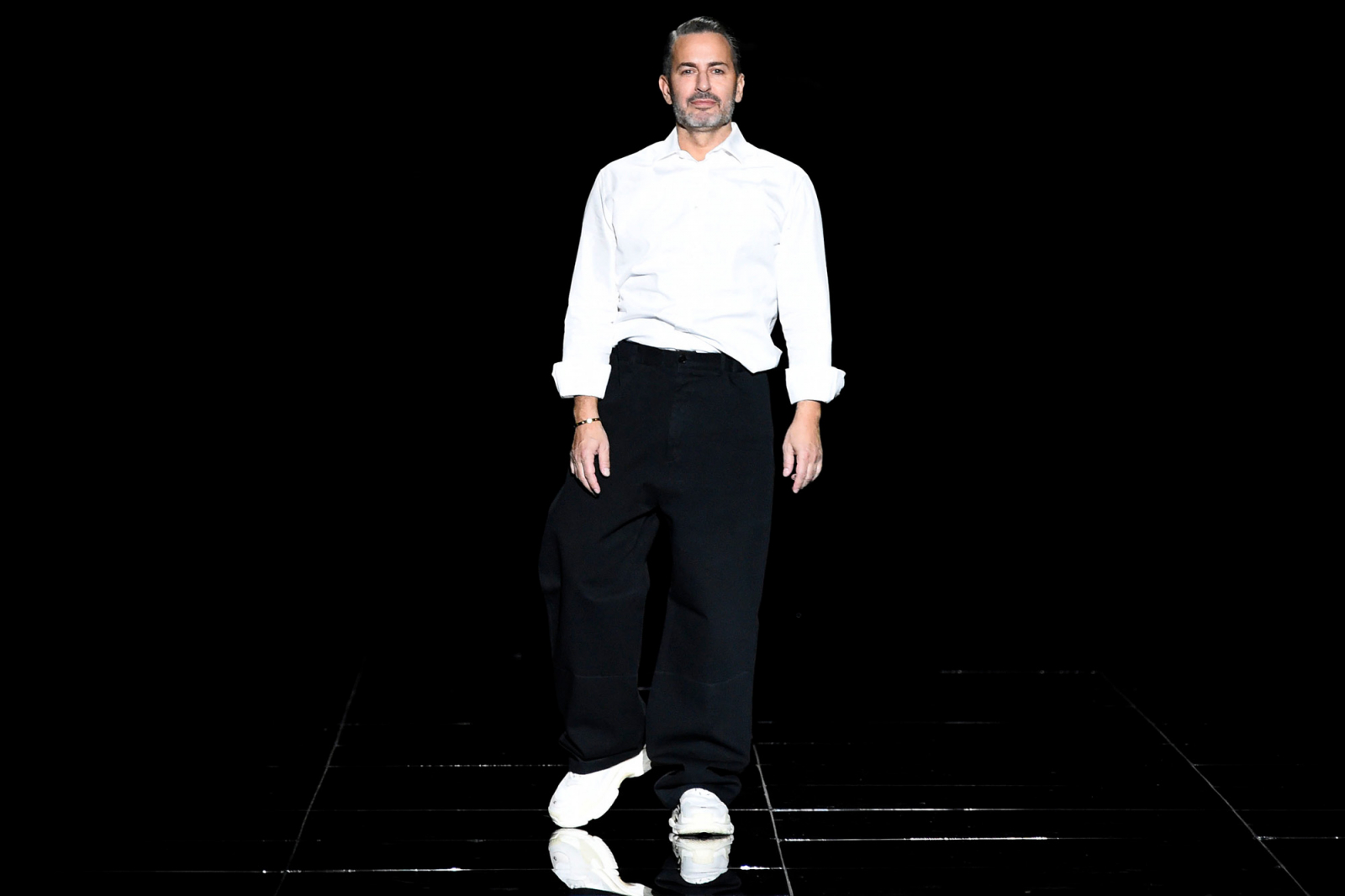 Marc Jacobs w białej koszuli i czarnych szerokich spodniach, stoi na wybiegu z czarnym tłem.