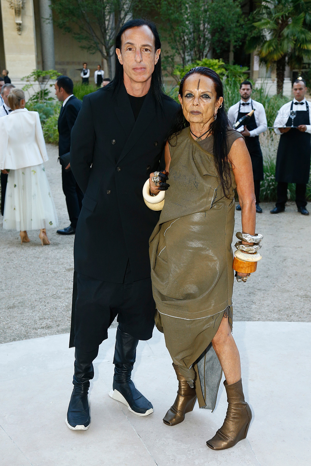 Rick Owens i Michele Lamy pozują w eleganckim ogrodzie, oboje w awangardowych stylizacjach.