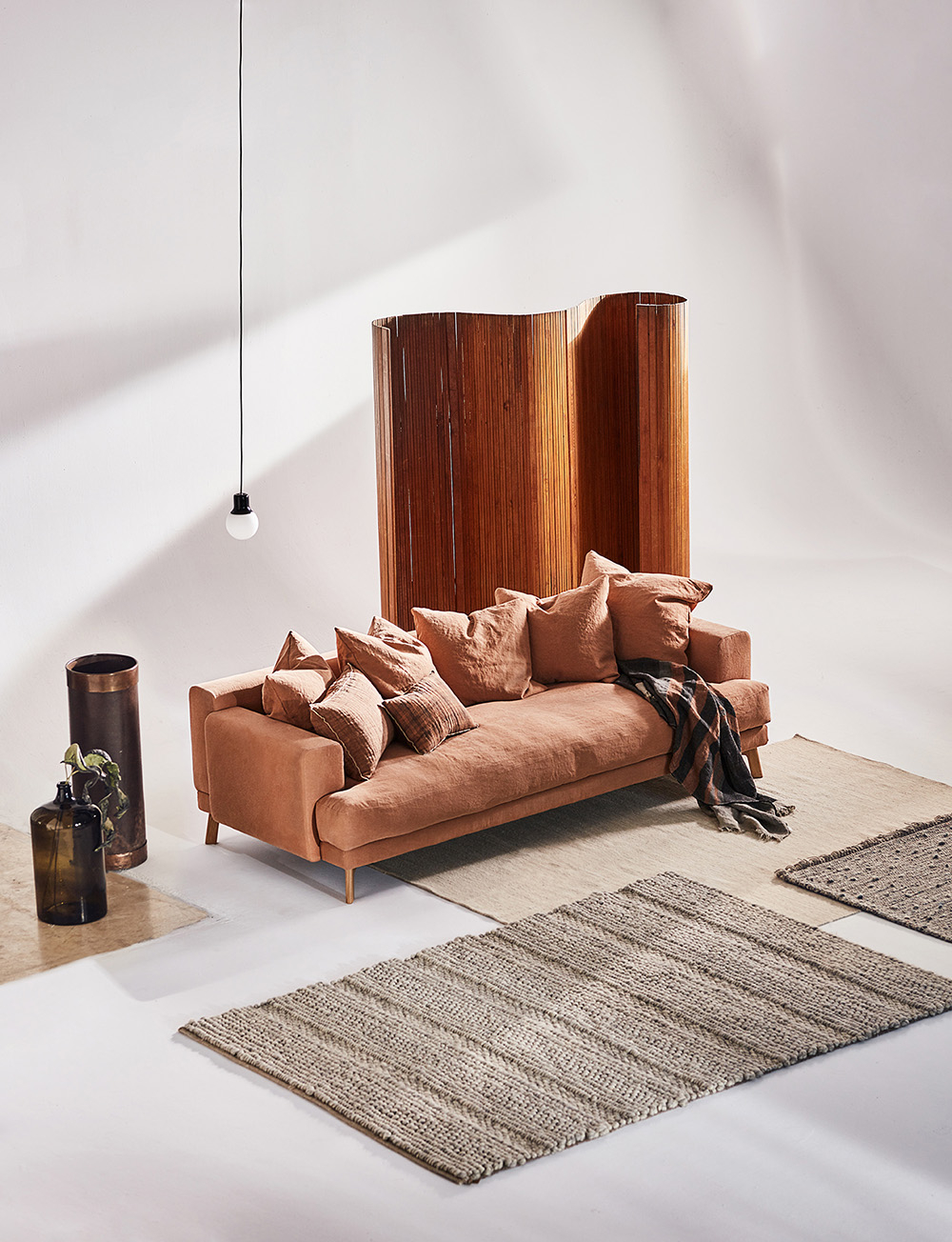 Różowa sofa z poduszkami, drewniana ścianka, lampa wisząca i dywany w stonowanych kolorach na jasnym tle.