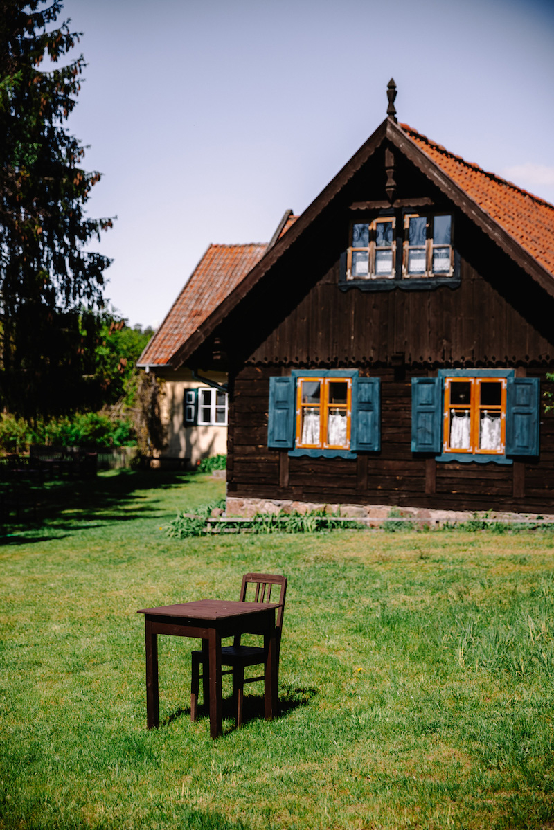 Na zielonym trawniku stoi stół z krzesłem, w tle widoczny drewniany dom z niebieskimi oknami i czerwoną dachówką.