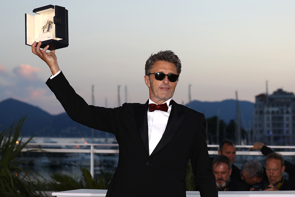 Paweł Pawlikowski trzyma w dłoni nagrodę, uśmiechając się i nosząc elegancki garnitur oraz okulary przeciwsłoneczne.