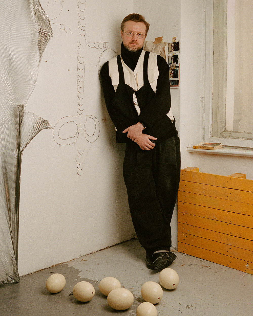 Piotr Bury Łakomy stoi w atelier, oparty o ścianę z rysunkiem. W tle leżą jaja i drewniane skrzynki.
