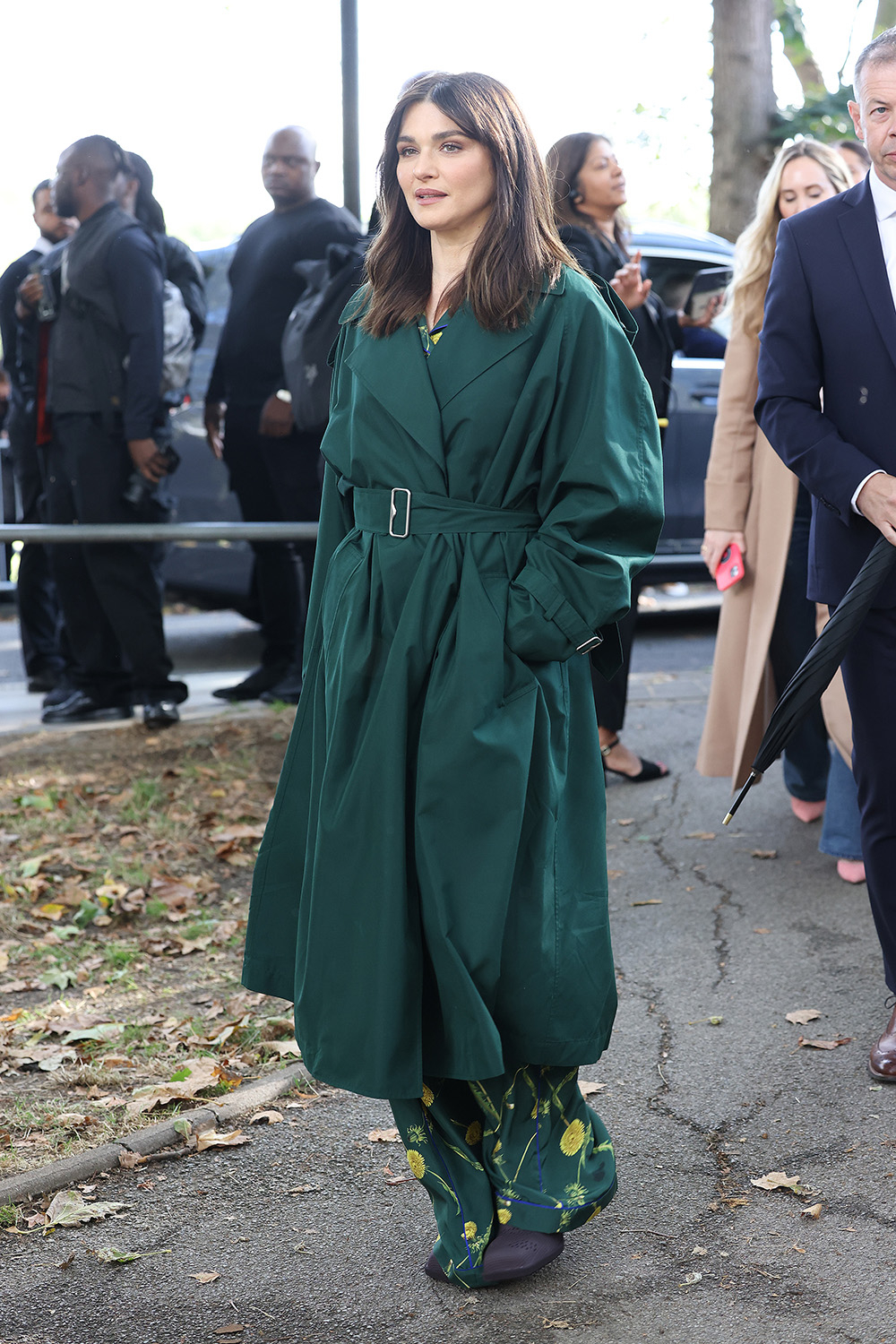 Rachel Weisz w zielonym trenczu Burberry, spacerująca wśród ludzi na ulicy, w towarzystwie innych osób.