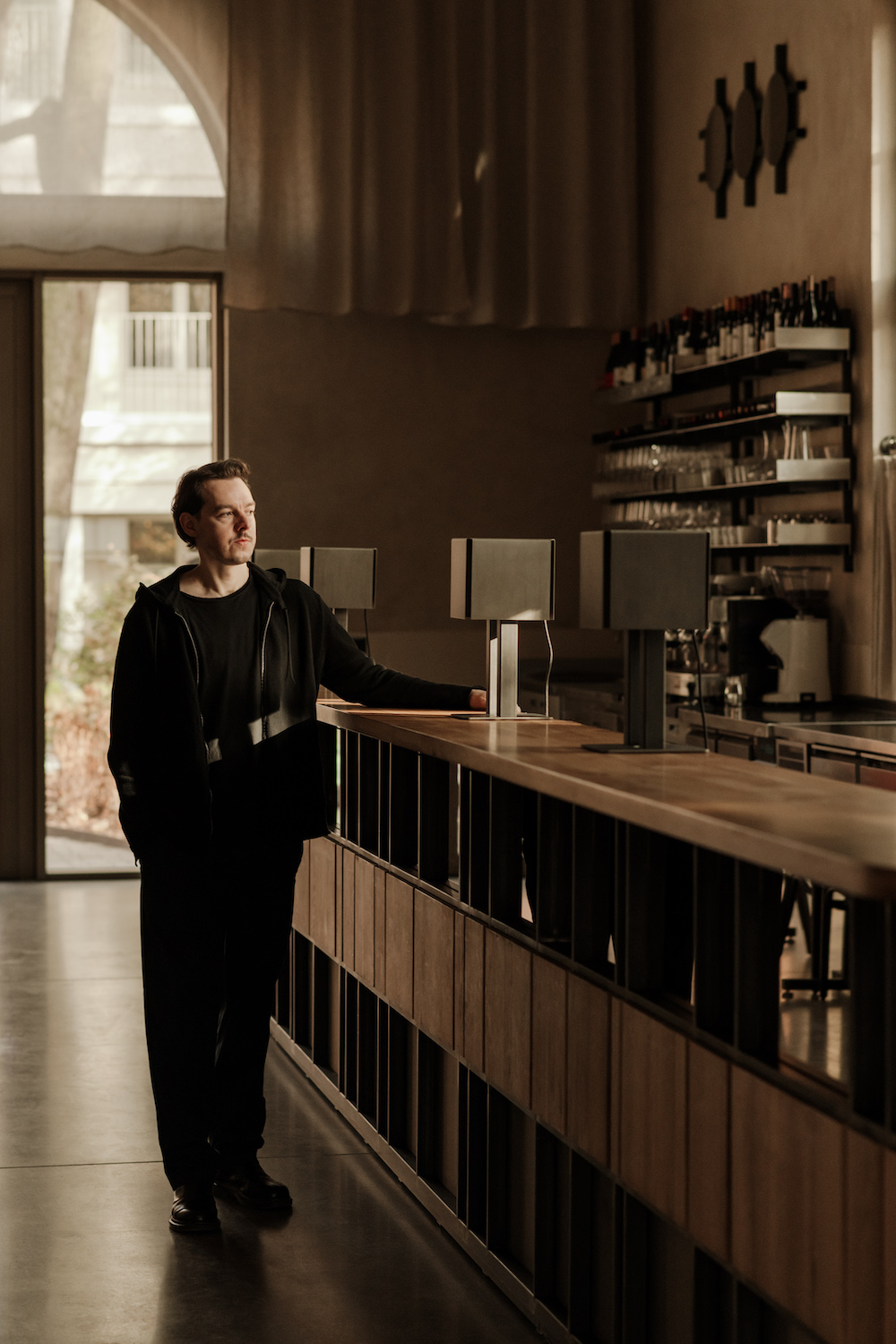 Adam Wierciński stoi przy barze w stylowej kawiarni, w tle widoczne półki z winem i lampy.