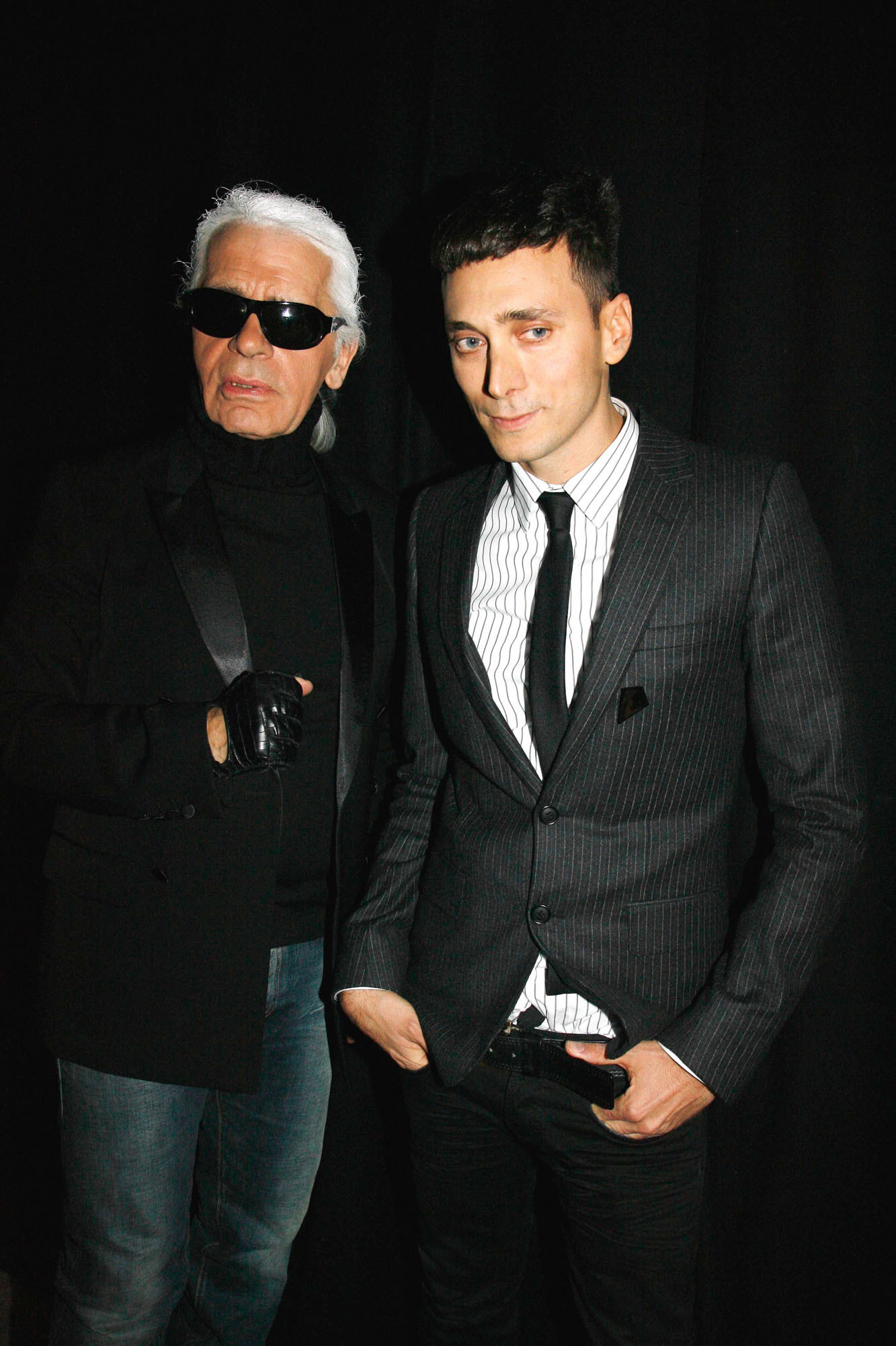 Karl Lagerfeld i Hedi Slimane w eleganckich strojach, pozujący na tle czarnej ściany.
