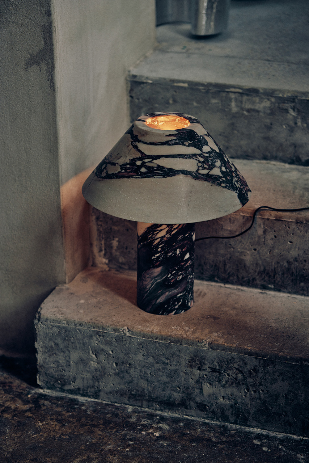 Lampa stojąca z betonowym kloszem i kolorowym podstawą, ustawiona na schodach o surowym wykończeniu.