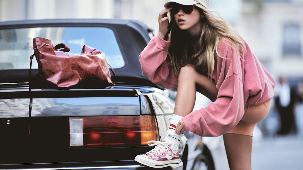 Lila Moss w różowej bluzie i czapce, zakłada sneakersy przy czarnym samochodzie. Modna stylizacja, torba na tle.