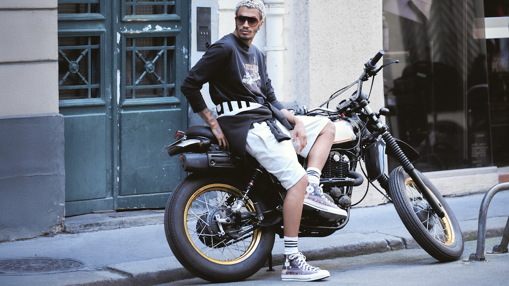Mężczyzna z blond włosami siedzi na czarnej motocyklu, ubrany w luźną bluzę i szorty, w tle zabytkowe drzwi.