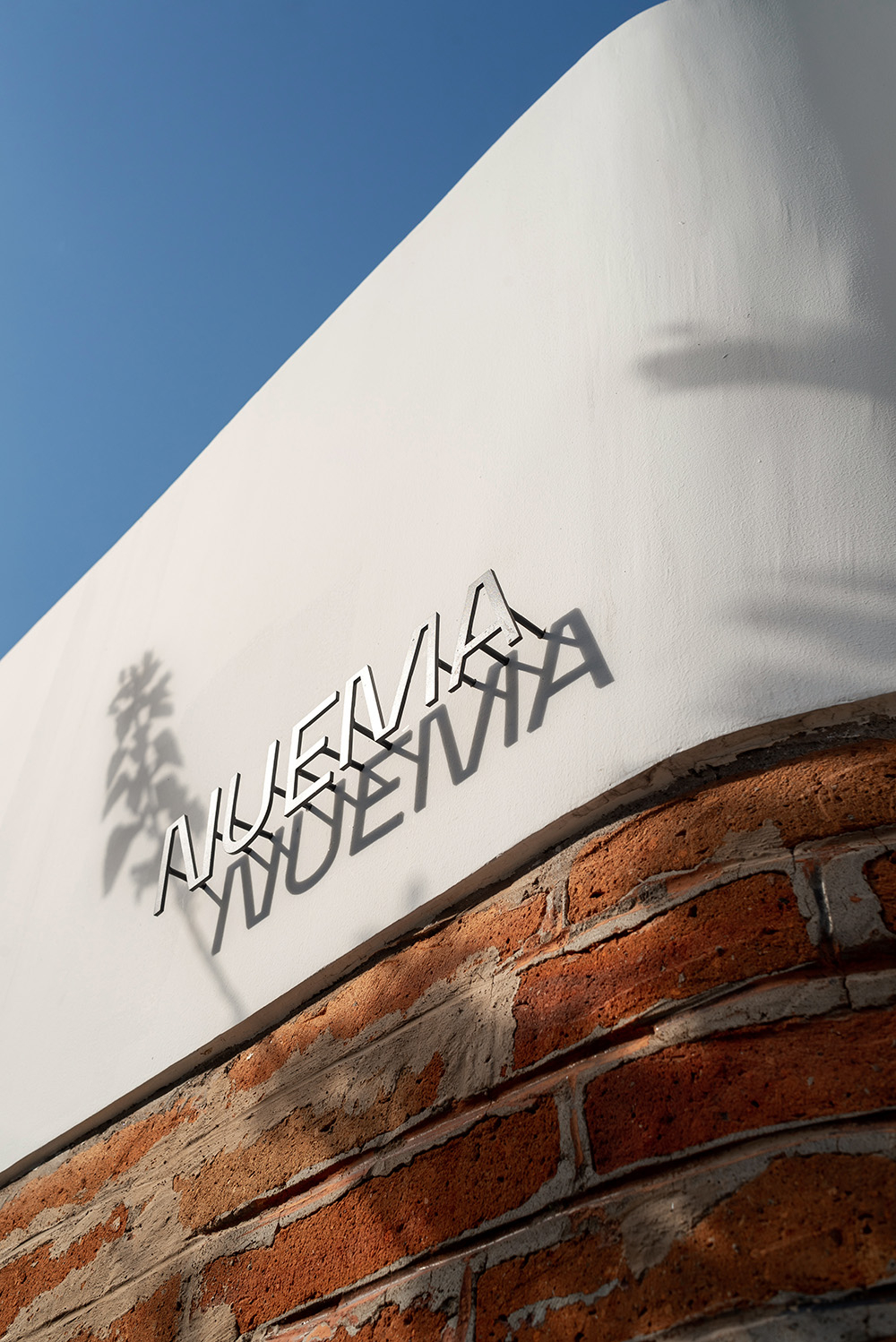 Na białej ścianie znajduje się napis "Nuevía", z cieniami i fragmentem ceglanej ściany. Tło jest błękitne.