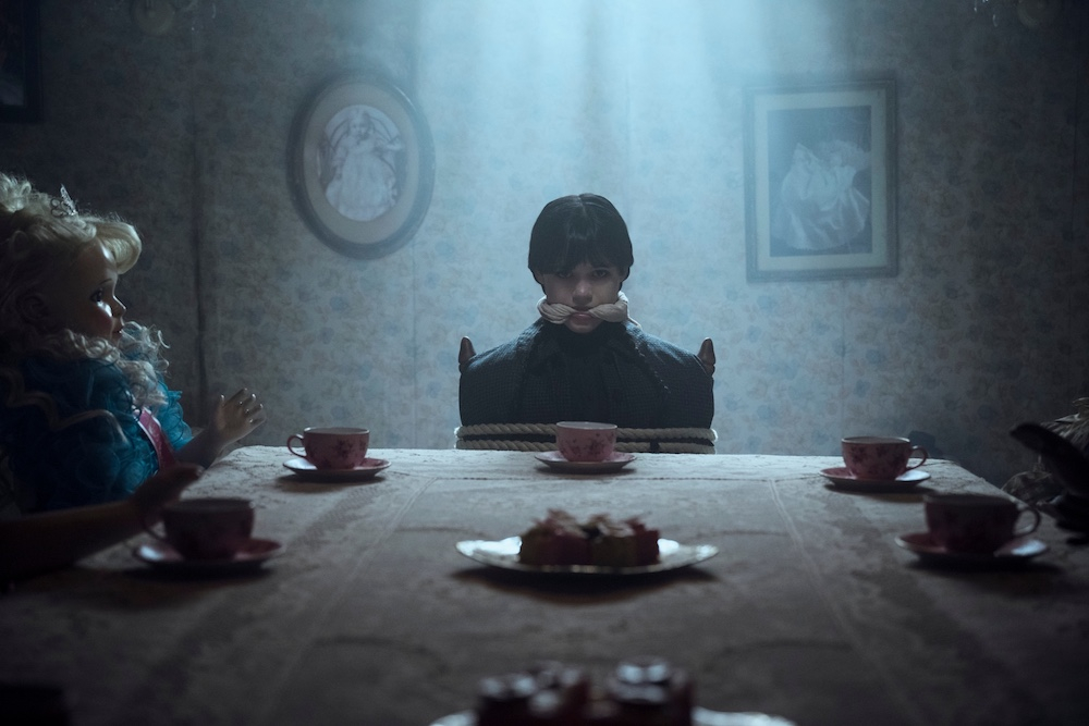 Chłopiec przebrany za postać z serialu "Wednesday" Netflix siedzi przy stole z lalkami, w mrocznym otoczeniu.