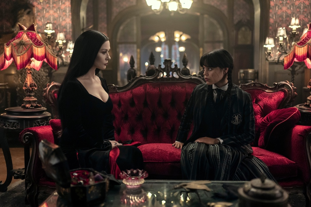 Dwóch bohaterów z serialu "Wednesday" na Netflixie siedzi na czerwonym drewnianym sofie w eleganckim otoczeniu.