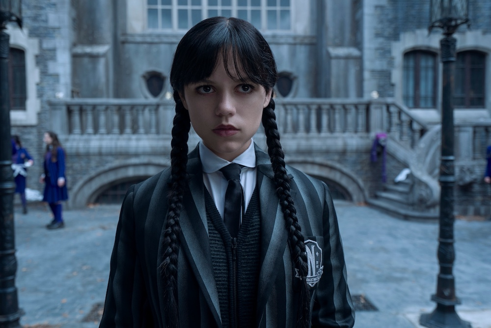 Postać z serialu "Wednesday" na Netfliksie, ubrana w czarny garnitur, stoi przed budynkiem.