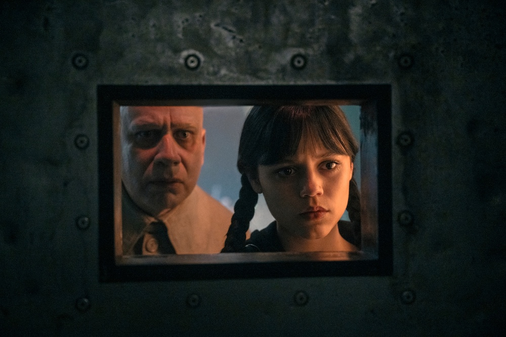 Kadr z serialu "Wednesday" na Netflix przedstawia postacie obserwujące przez metalowe okienko.