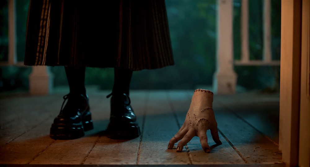Ręka-figuryna leży na podłodze, obok postaci w stylowych butach. Scena inspirowana serialem "Wednesday" na Netflixie.