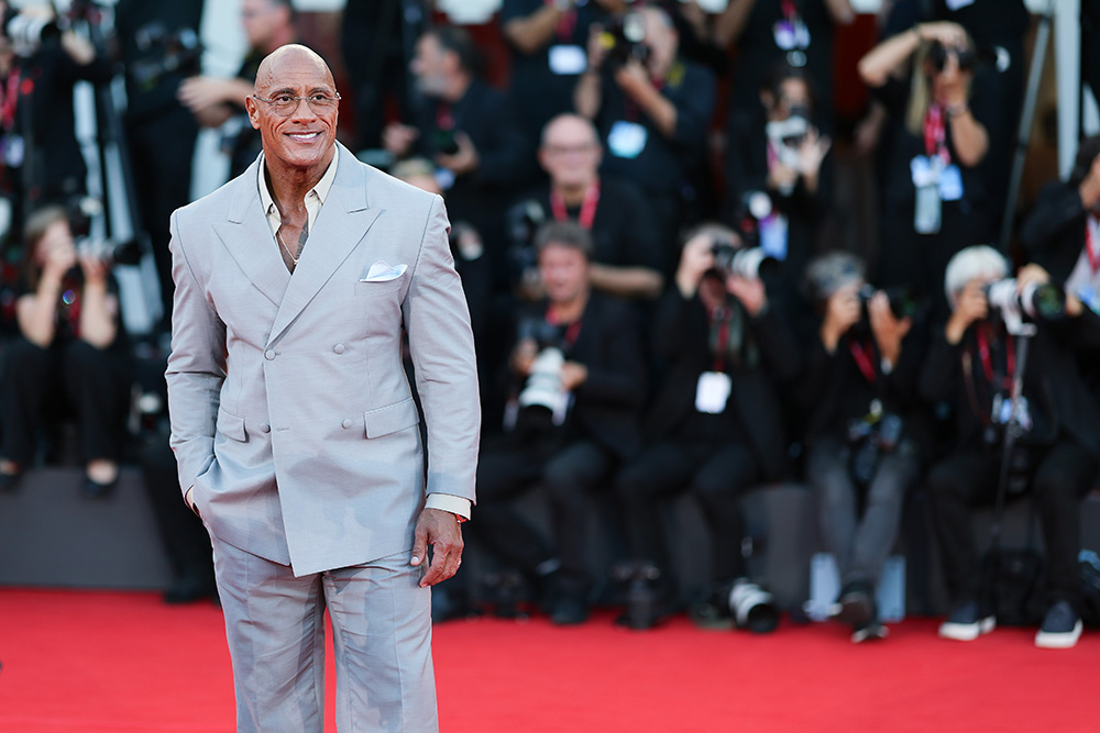 Dwayne Johnson w eleganckim szarym garniturze na czerwonym dywanie, otoczony przez fotografów.