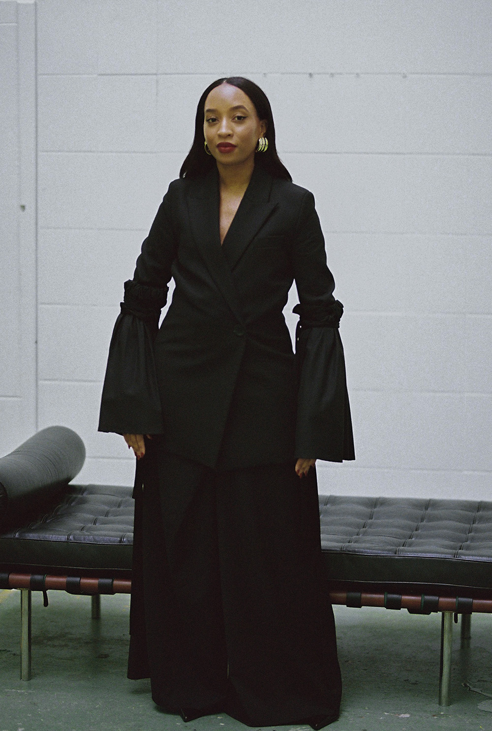 Stephany Goncalves stoi w czarnym eleganckim stroju, z długimi rękawami, na tle białej ściany.