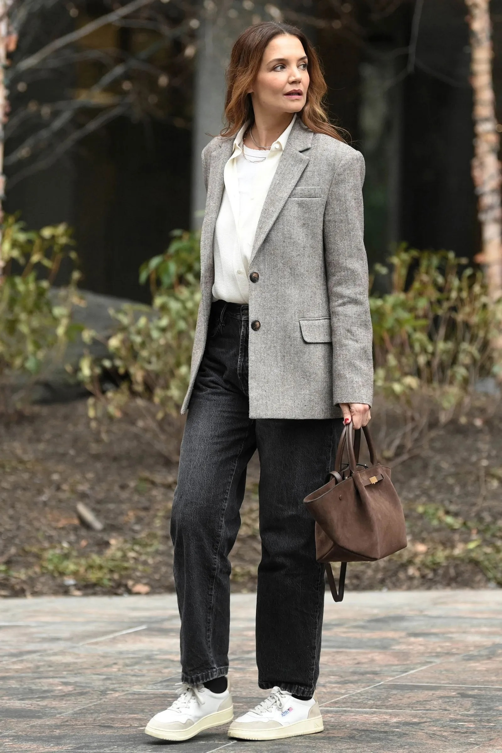 Katie Holmes w szarej marynarce, czarnych spodniach i sneakersach Autry, trzymając małą brązową torebkę.