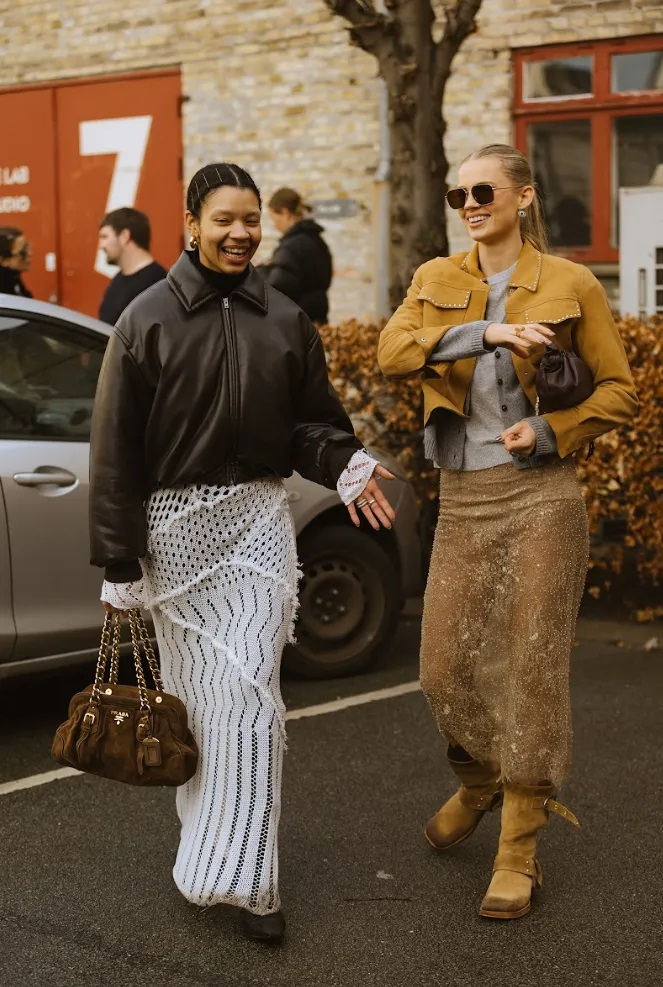 Dwie kobiety w modnych strojach, jedna w spódnicy boho z drapowaniem, druga w eleganckim zestawie z kurtką.