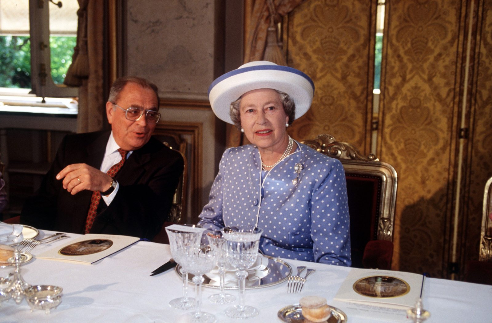 Królowa Elżbieta II w niebieskiej sukience w kropki, z kapeluszem, podczas eleganckiego przyjęcia.