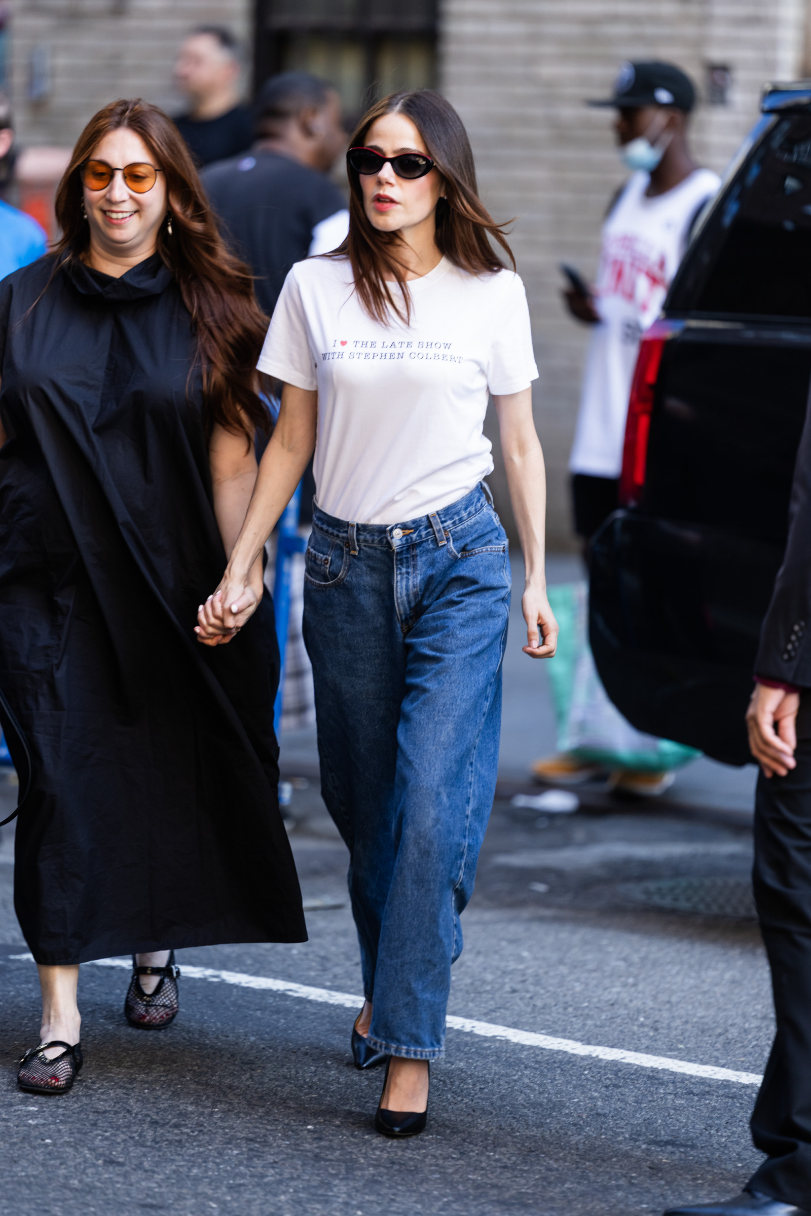 Kobieta w białym T-shirtcie i jeansach spaceruje ulicą, trzymając się za ręce z inną osobą.