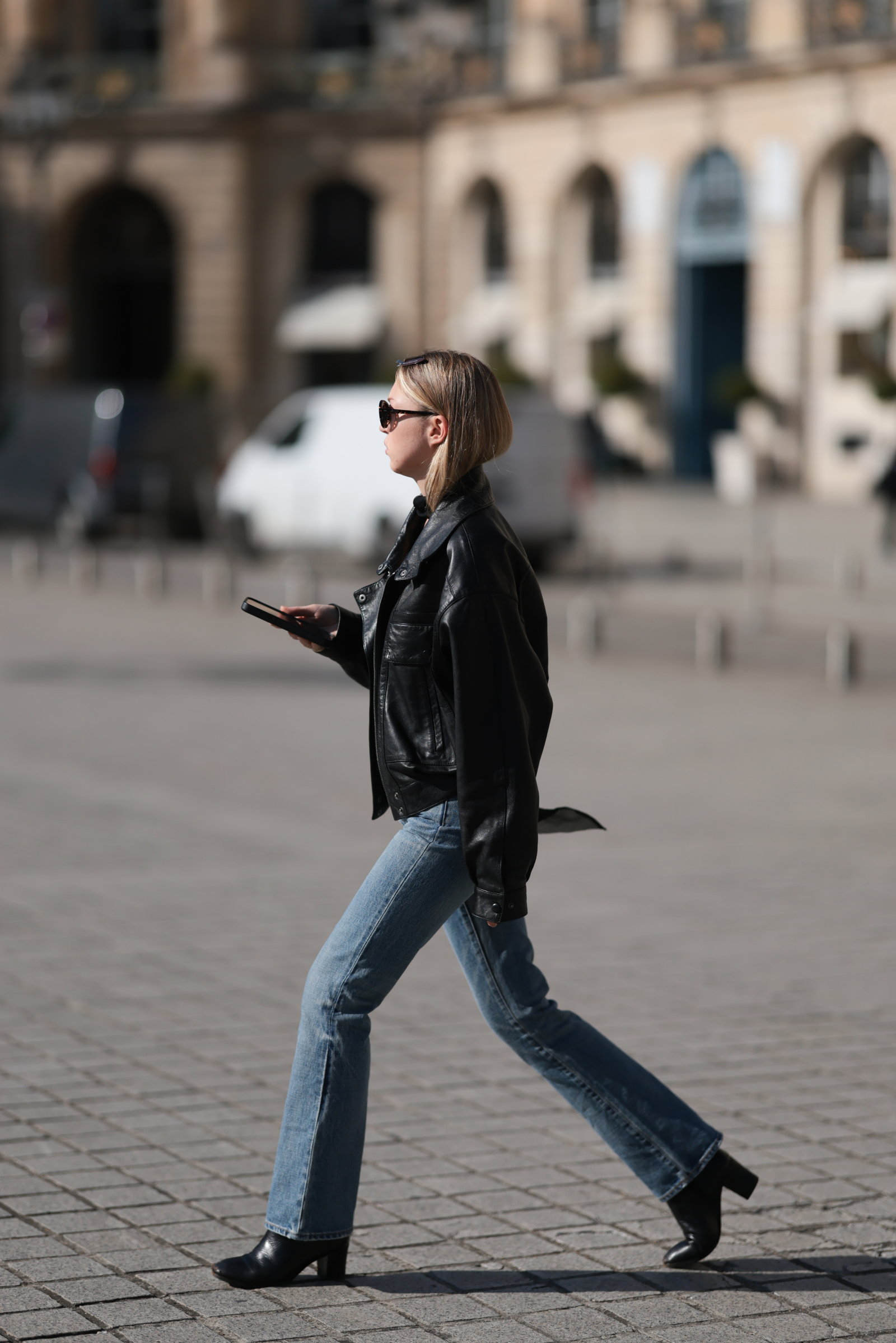Kobieta w skórzanej kurtce, jeansach bootcut i czarnych botkach, spacerująca z telefonem w dłoni.