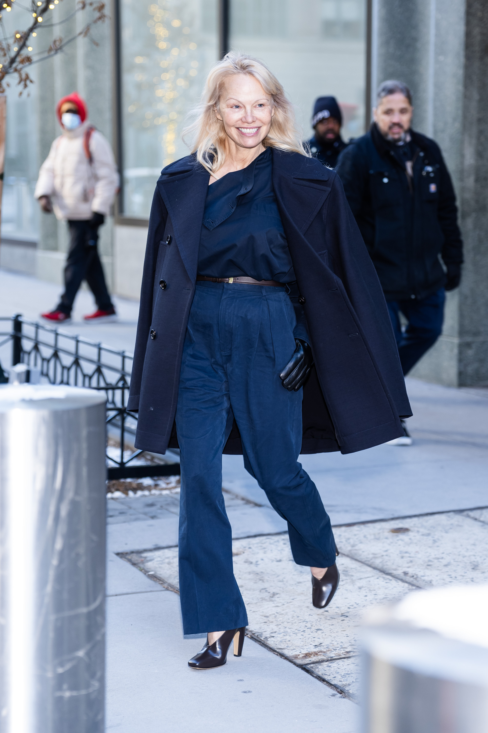 Pamela Anderson w ciemnej stylizacji, uśmiechając się, spaceruje po ulicy, w otoczeniu innych przechodniów.