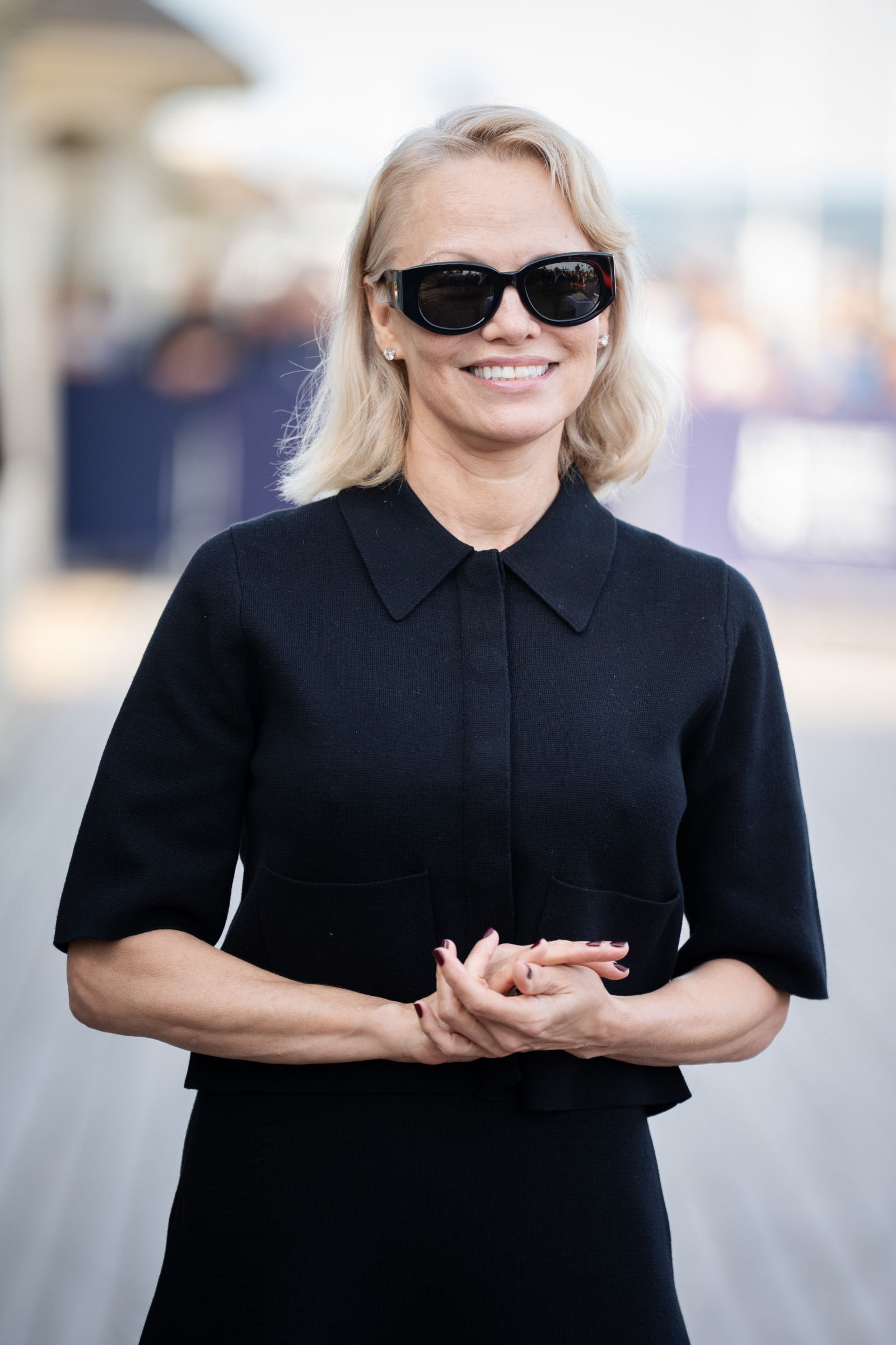 Pamela Anderson w eleganckim, czarnym kostiumie i okularach przeciwsłonecznych, uśmiechając się na tle wydarzenia.