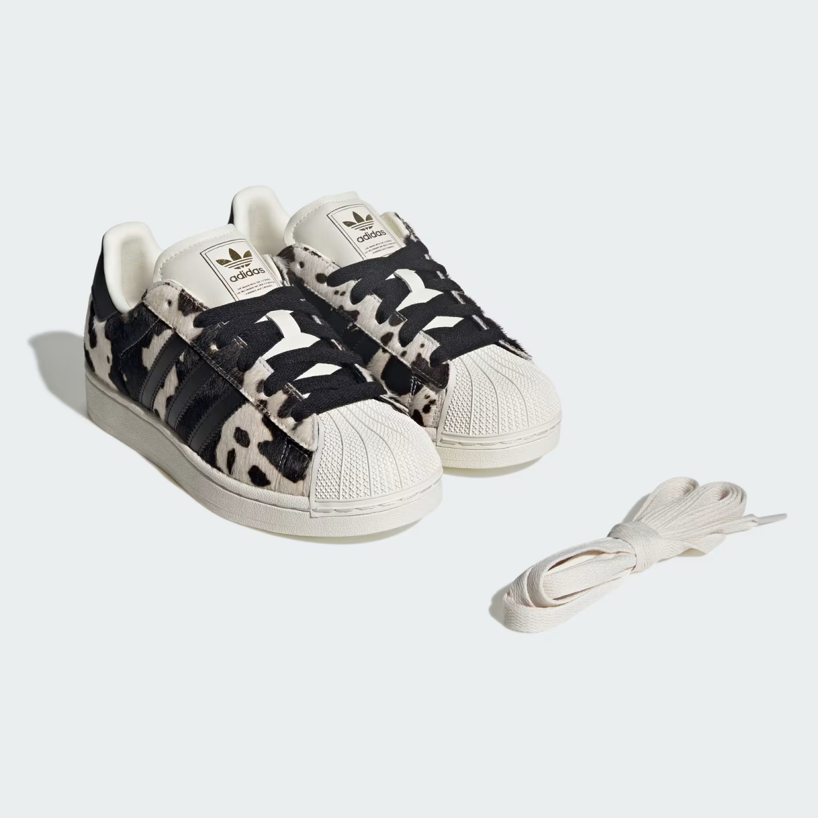 Sneakersy w krowie łaty, model adidas Superstar, z białą podeszwą i czarnym akcentem. Obok dodatkowe sznurówki.