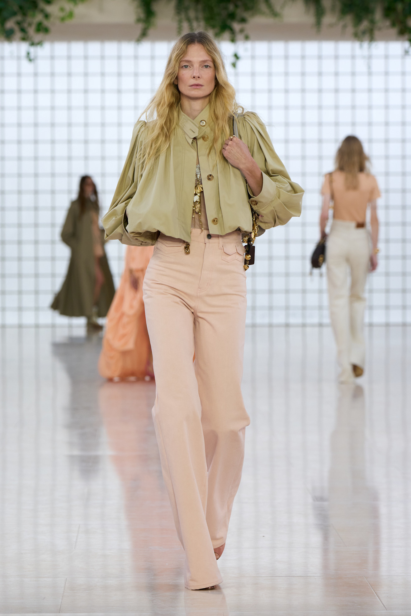 Modelka w różowych jeansach Chloe oraz oversize&#039;owej bluzie na wybiegu, w tle inne sylwetki.