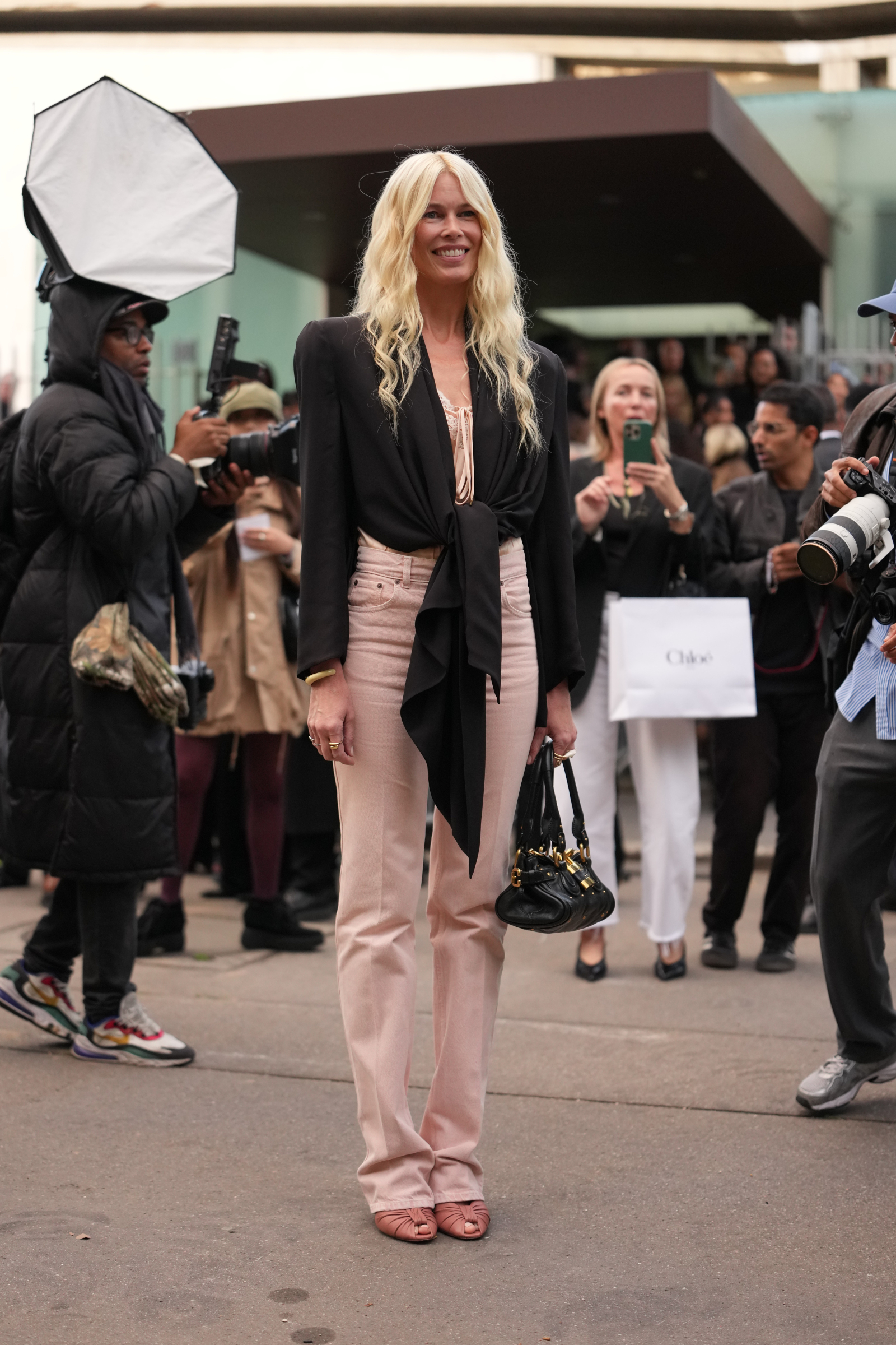 Claudia Schiffer w czarnym żakiecie i różowych jeansach, uśmiechnięta, na tle tłumu fotografów.