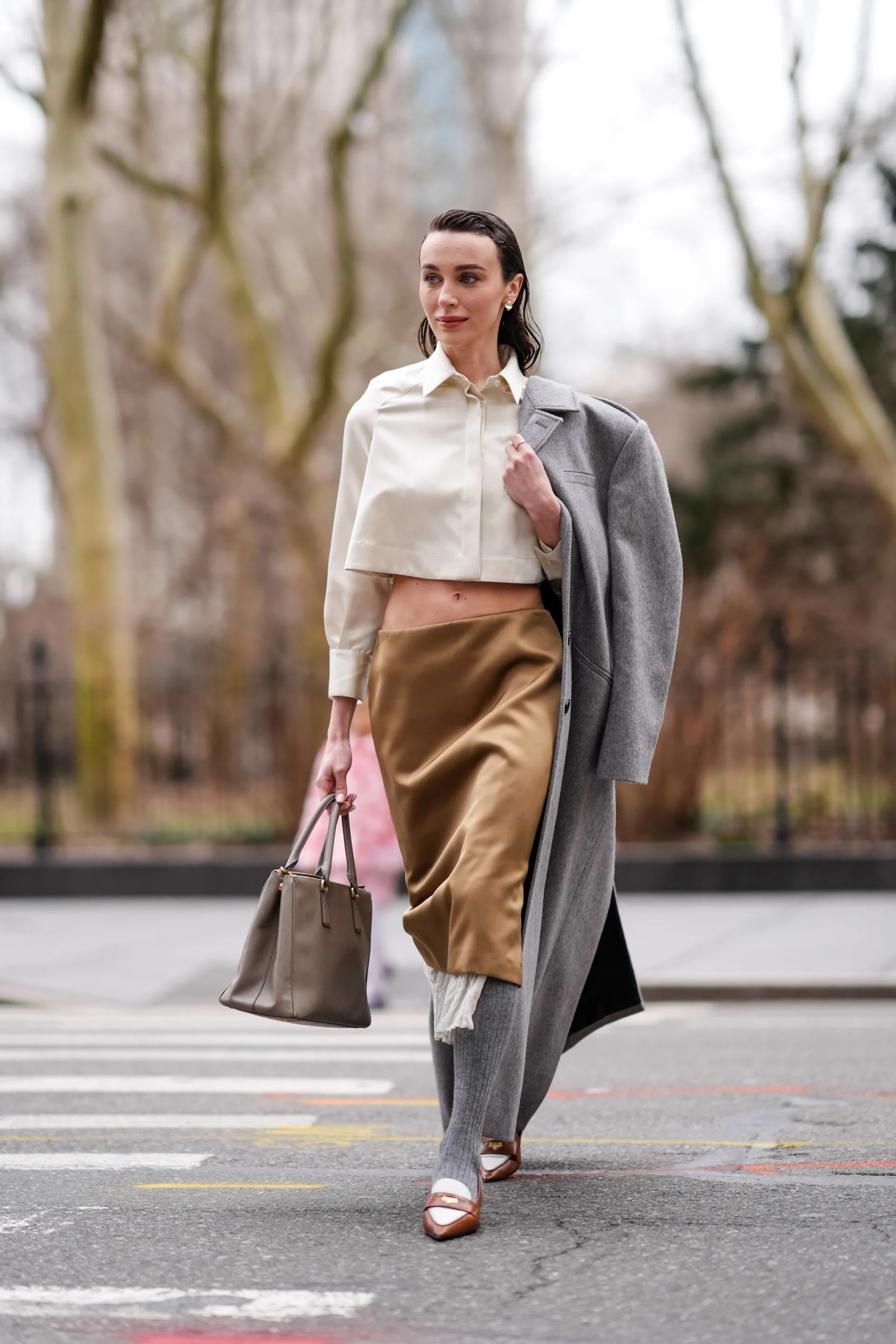 Kobieta idąca ulicą, ubrana w satynową spódnicę w kolorze złotym, białą bluzkę i szary płaszcz, trzyma torebkę.