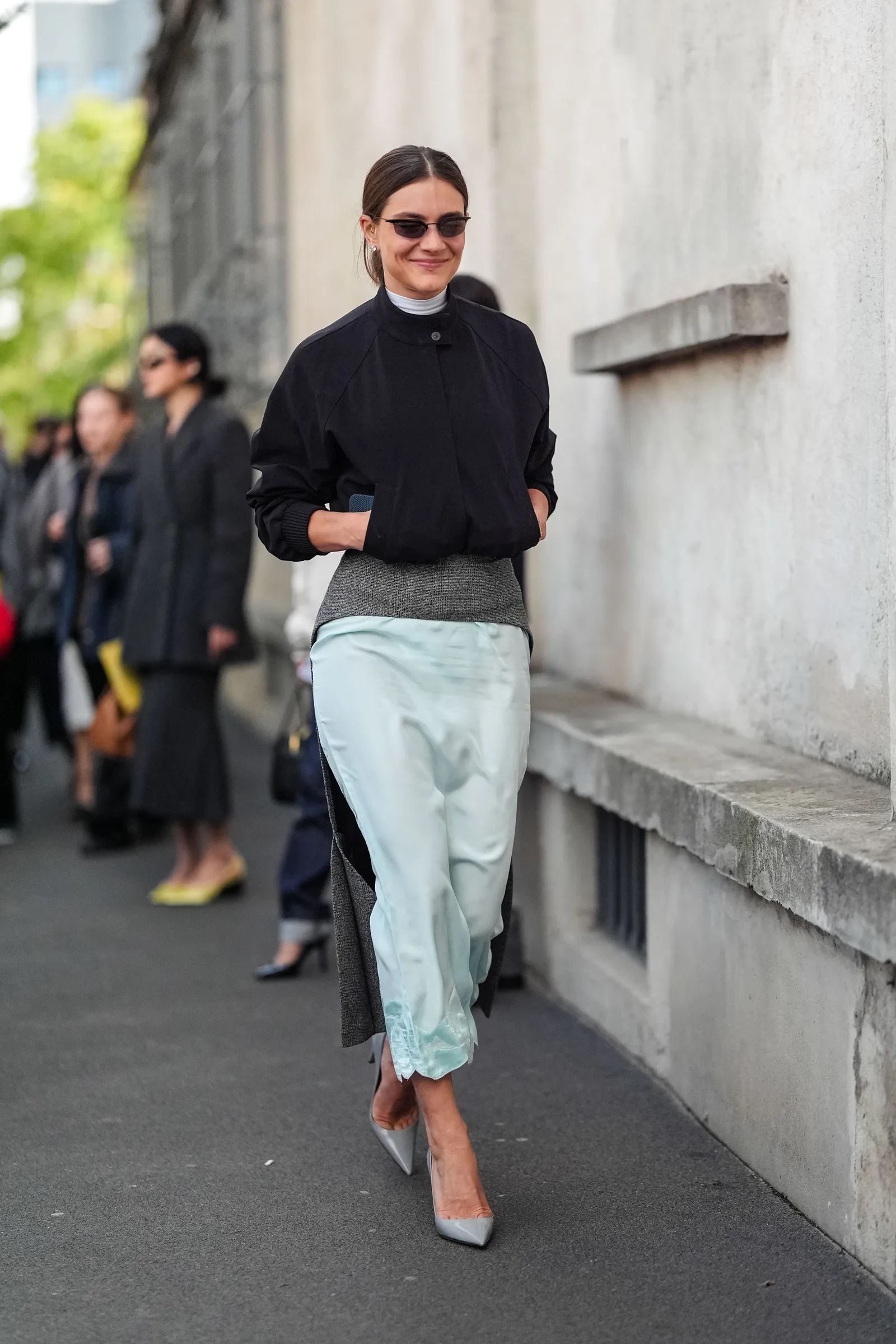 Kobieta w czarnej kurtce i satynowej spódnicy w kolorze miętowym spaceruje po ulicy.