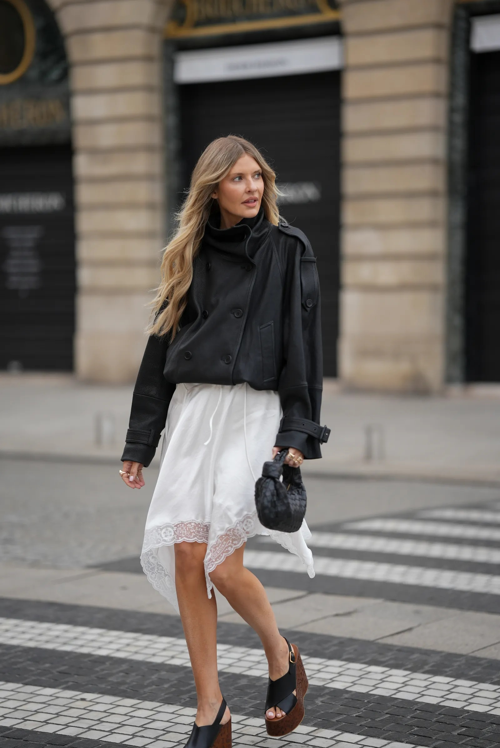 Kobieta w czarnej kurtce i satynowej spódnicy spaceruje po ulicy. Na stopach ma modne klapki.