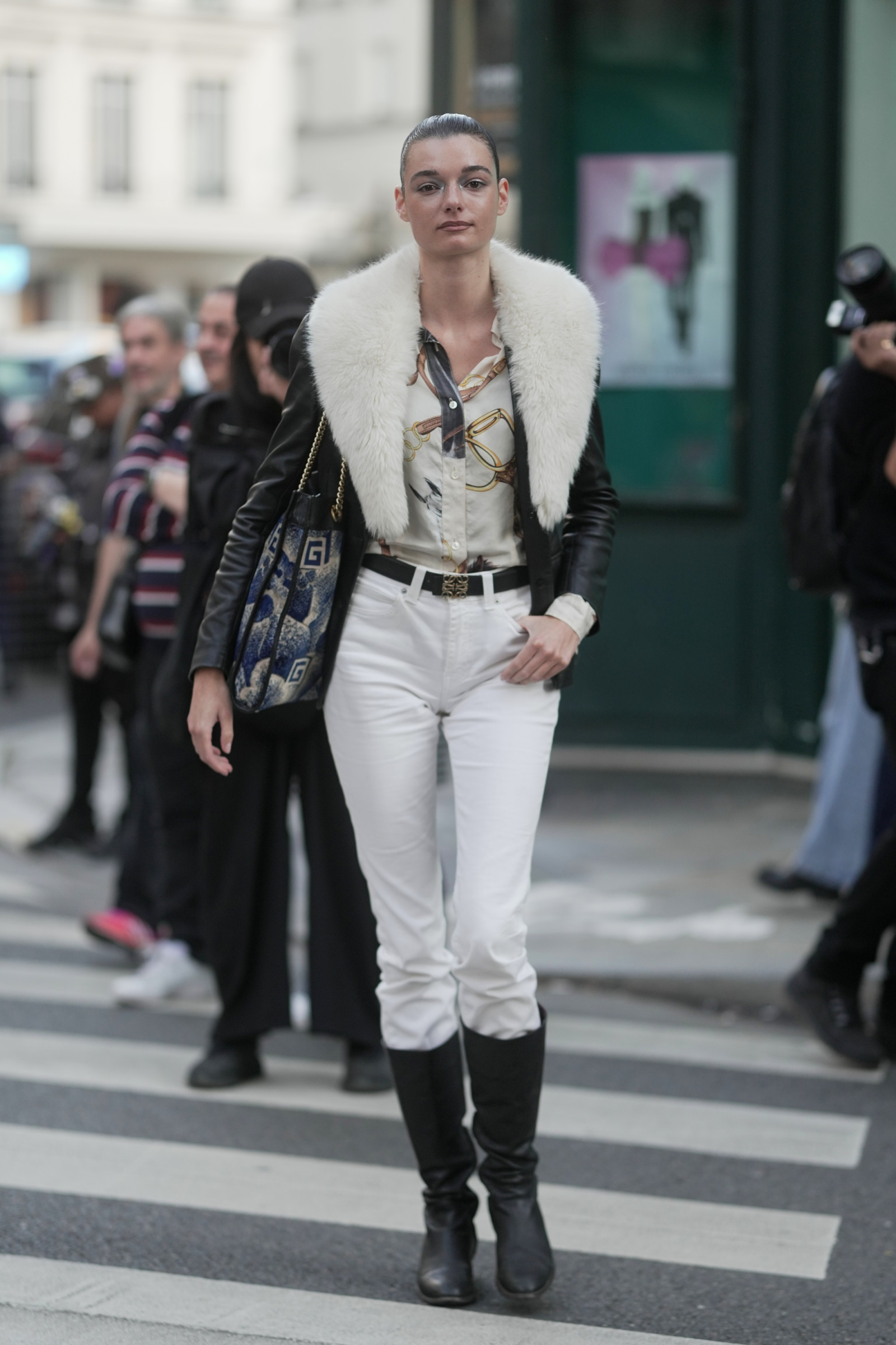 Kobieta w białych skinny jeans z czarnymi kozakami i futrzanym kołnierzem na kurtce, spacerująca po ulicy.