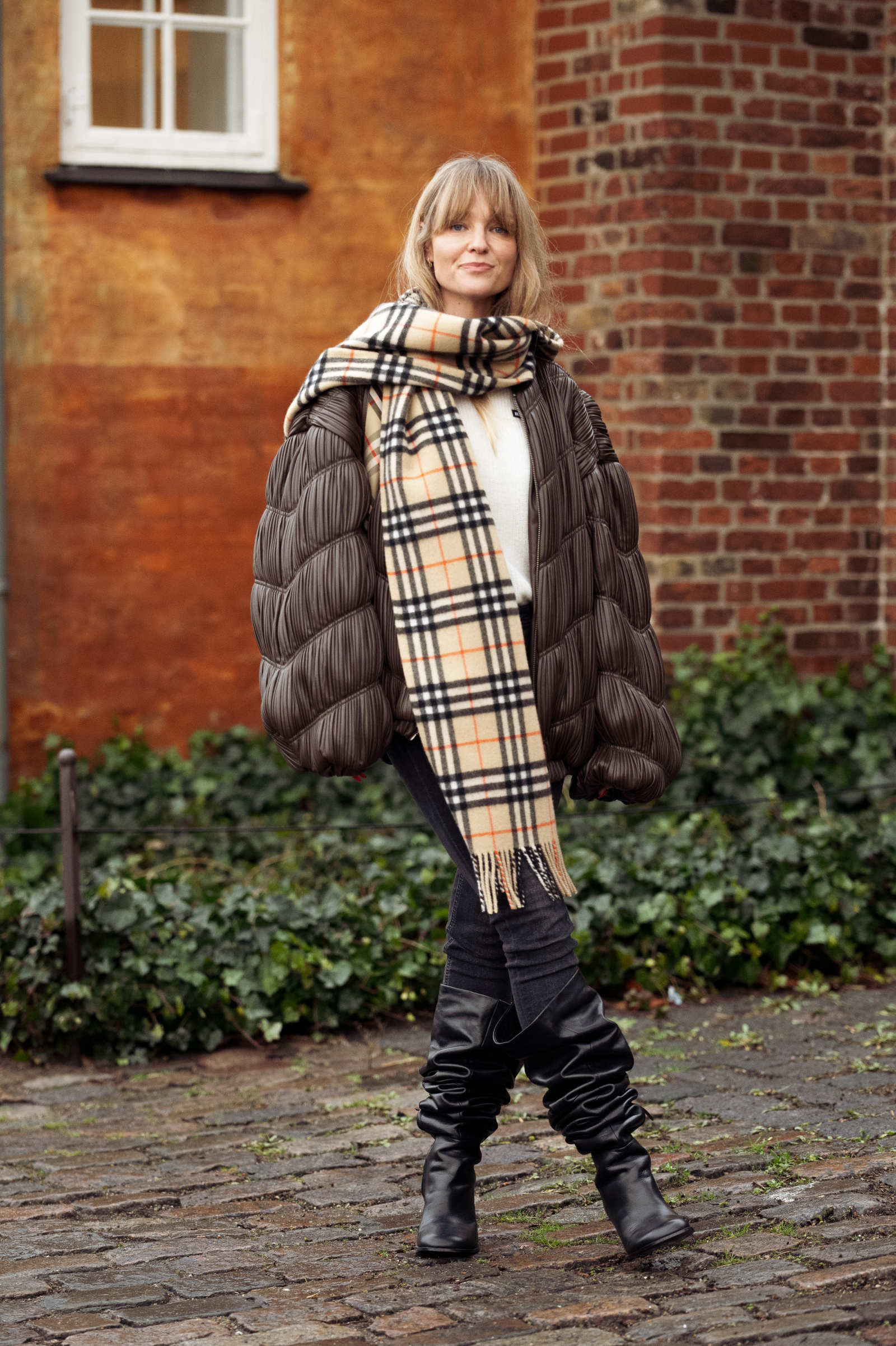 Kobieta w puchowej kurtce i zszarpanej chustce stoi na ulicy, nosząc skinny jeans i kozaki do kolan.