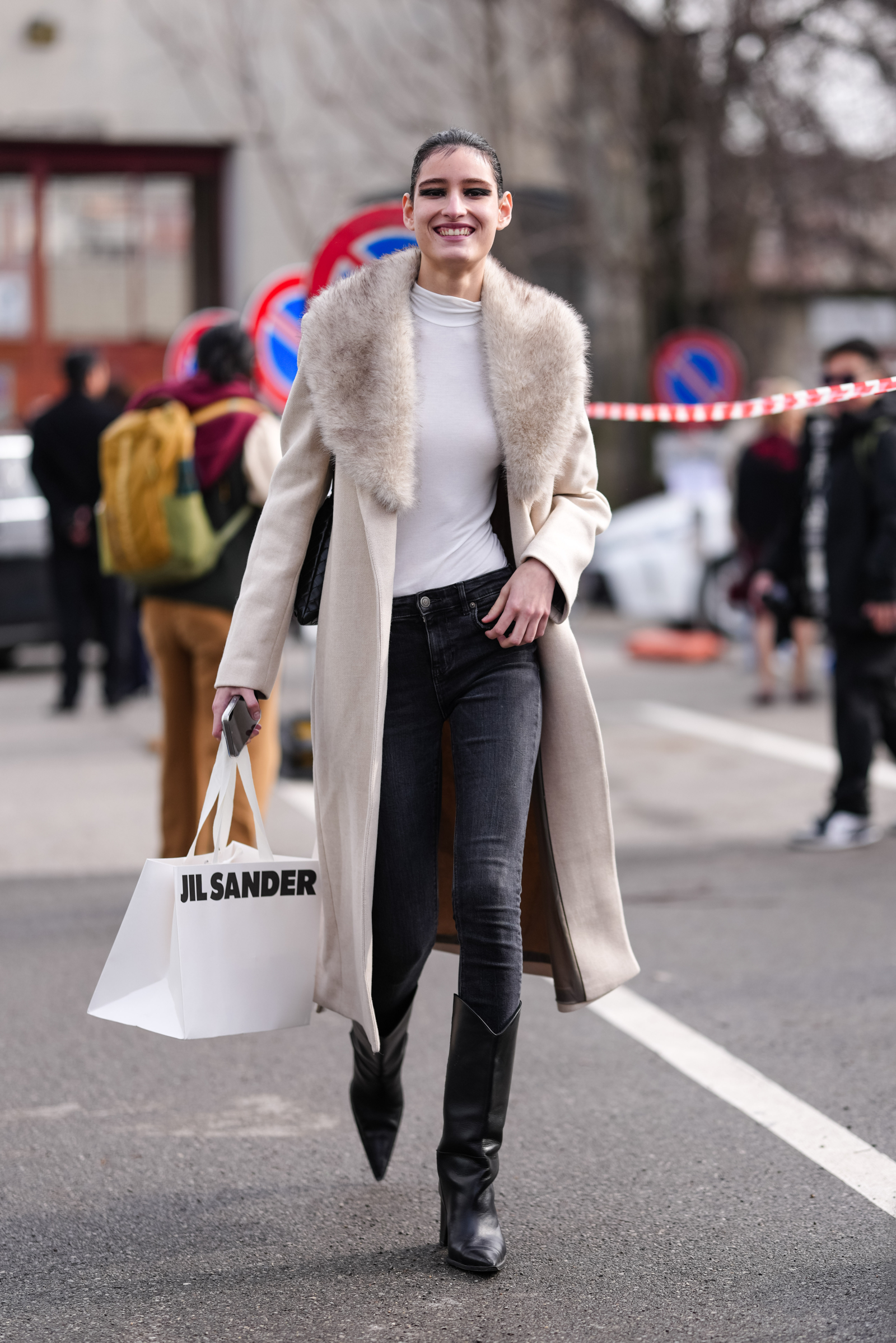 Kobieta w długim płaszczu z futrzanym kołnierzem, skinny jeans i kozaki, trzyma torbę z logo Jil Sander.