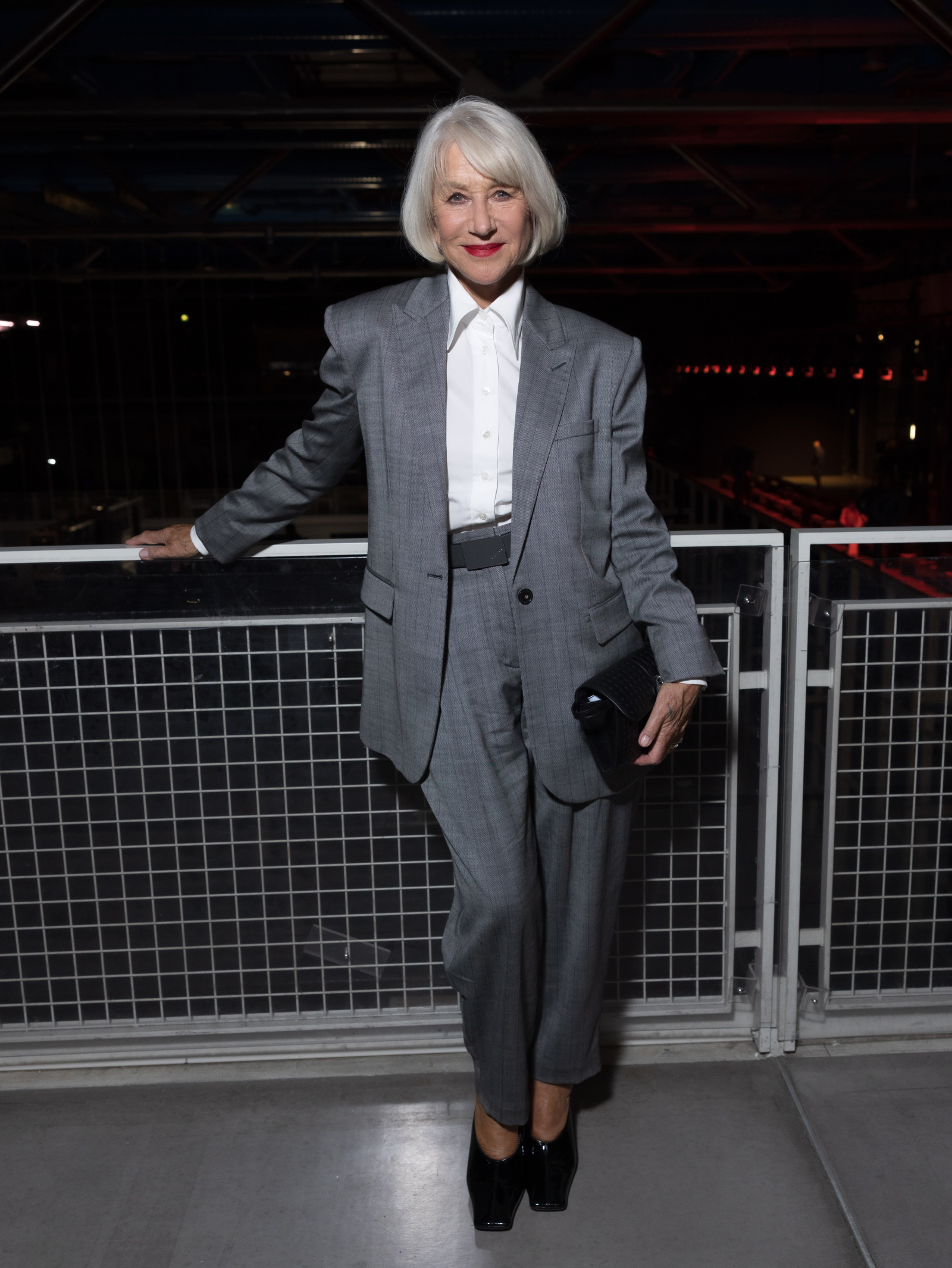 Helen Mirren w szarym garniturze, stojąca na tle nowoczesnego wnętrza, z torbą w ręku i uśmiechem na twarzy.