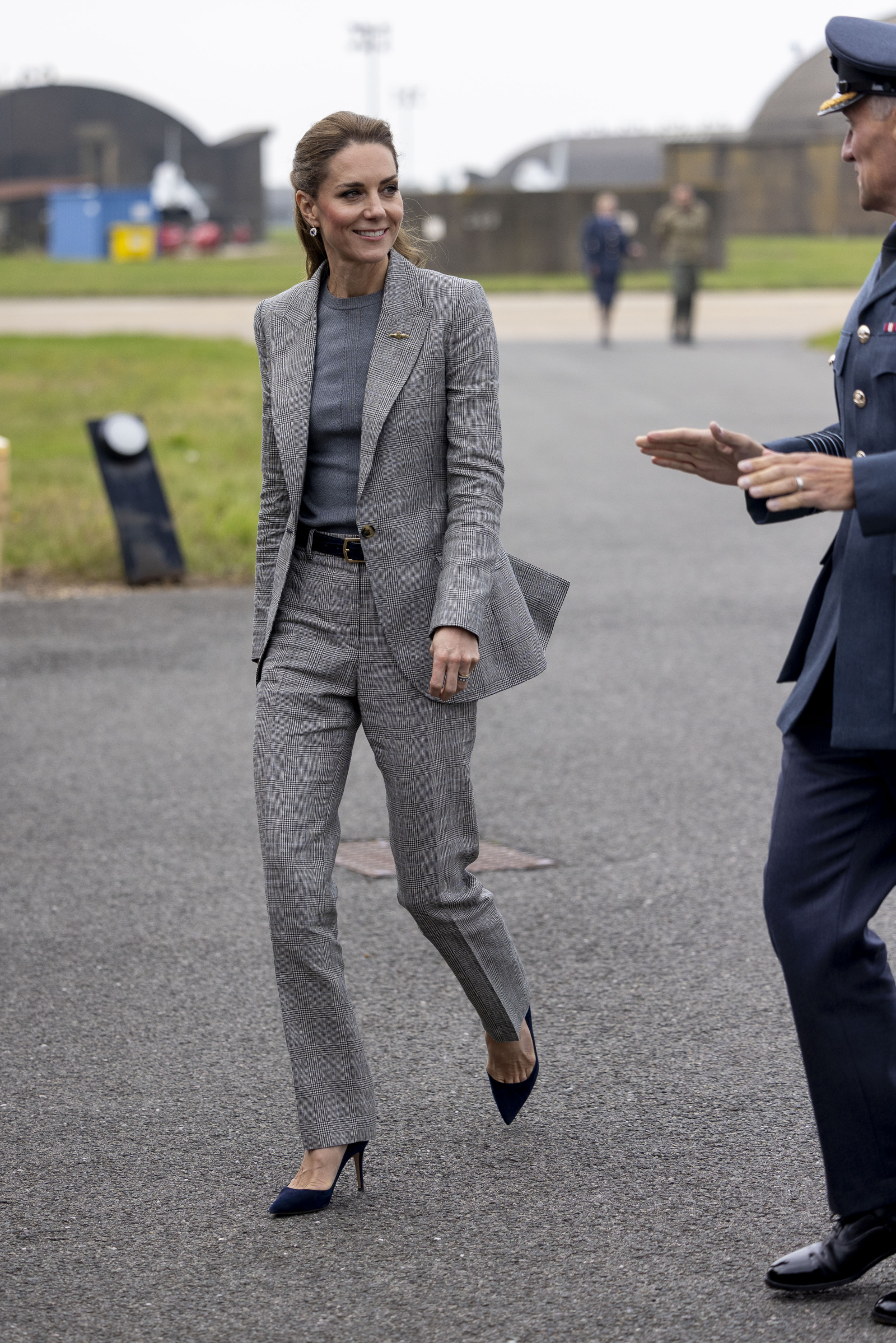 Księżna Kate w szarym garniturze spaceruje po terenie, rozmawiając z mężczyzną w mundurze.