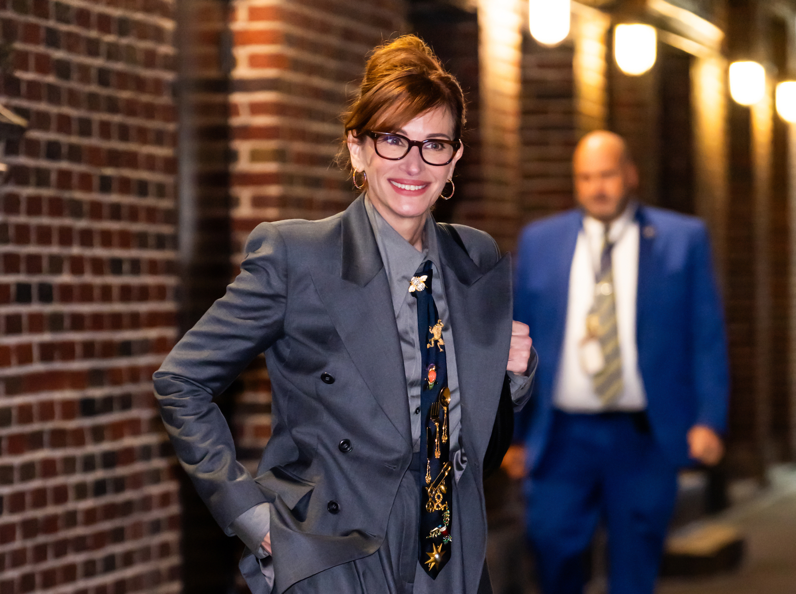 Julia Roberts w szarym garniturze, uśmiecha się, przechadzając się po oświetlonym korytarzu.