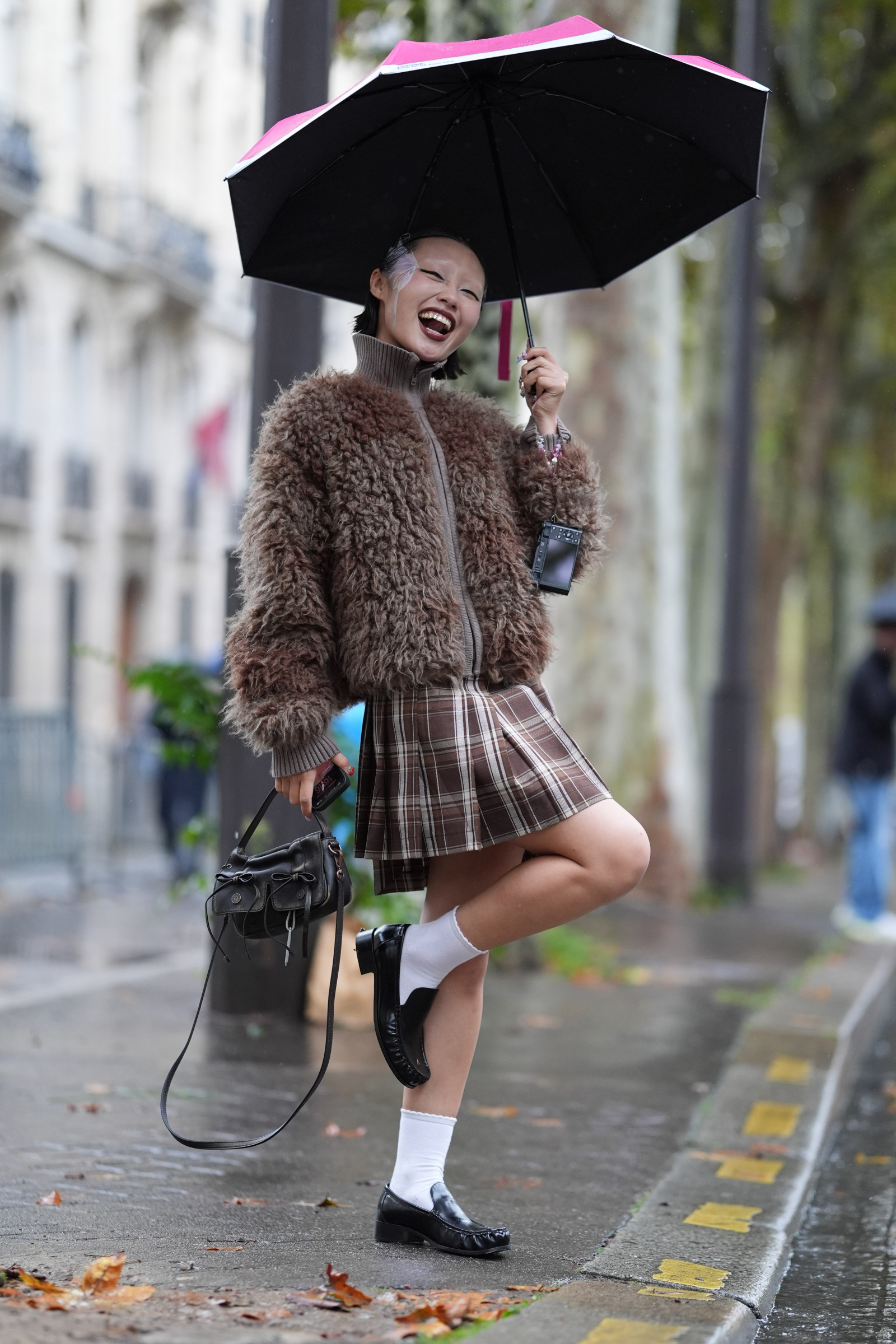 Kobieta w pluszowej kurtce teddy coat tańczy pod parasolem w deszczowej scenerii, ubrana w spódniczkę w kratę.