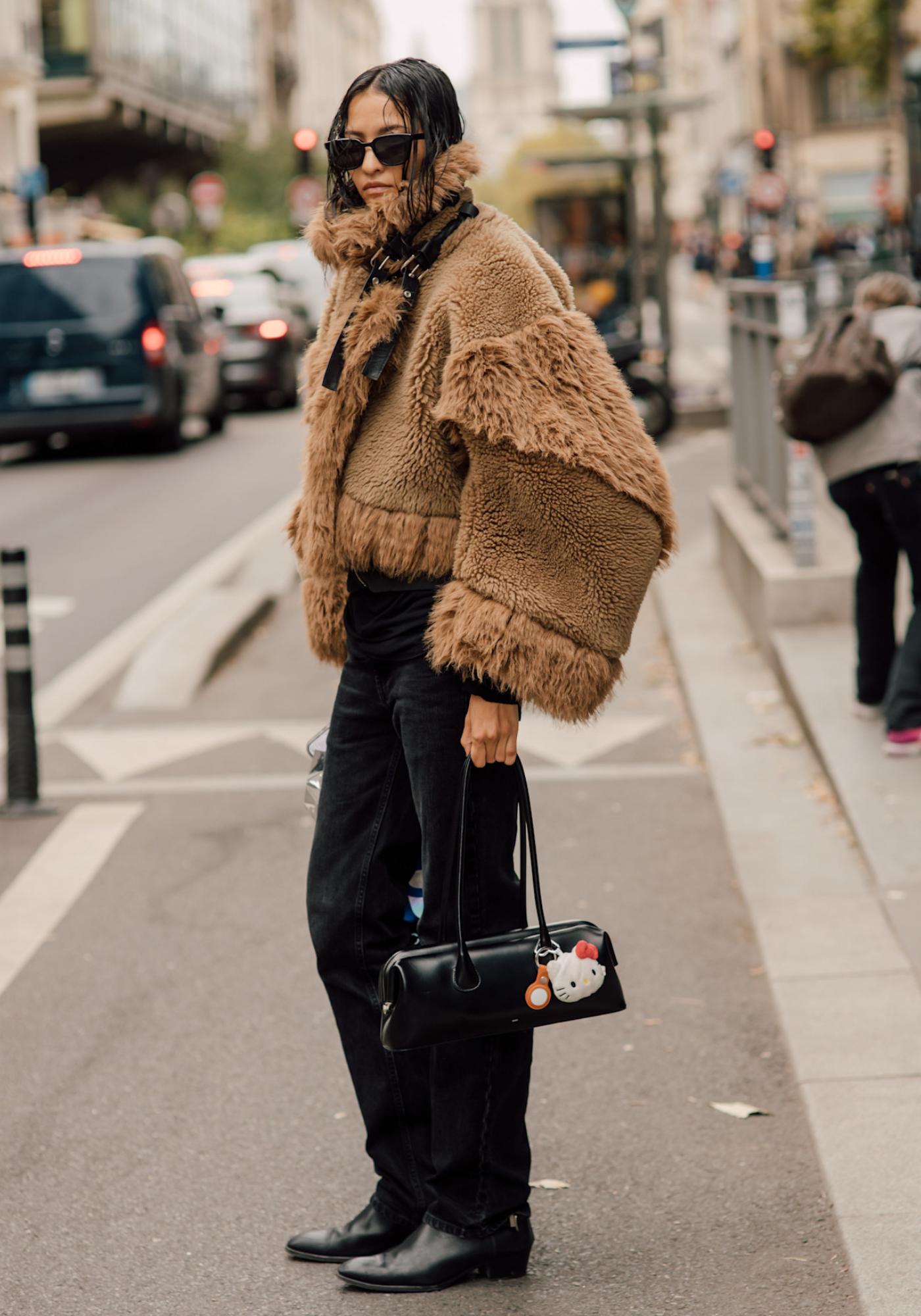 Osoba w ulicznej scenerii nosi kurtkę teddy coat w kolorze brązowym, czarne spodnie i torebkę.
