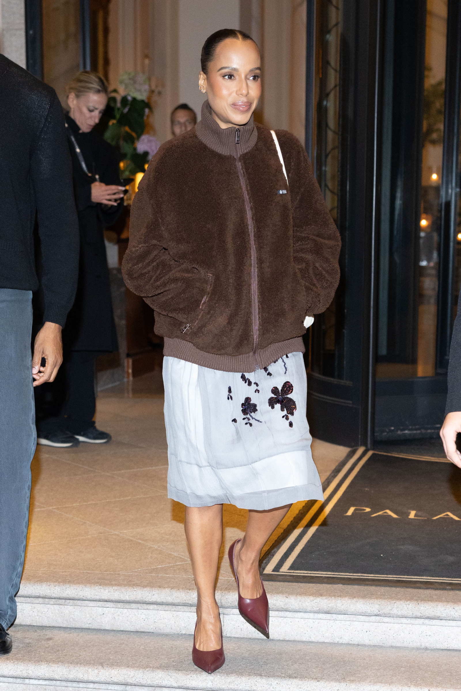 Kobieta wychodzi z budynku w brązowej kurtce teddy coat oraz eleganckiej spódnicy z haftem.