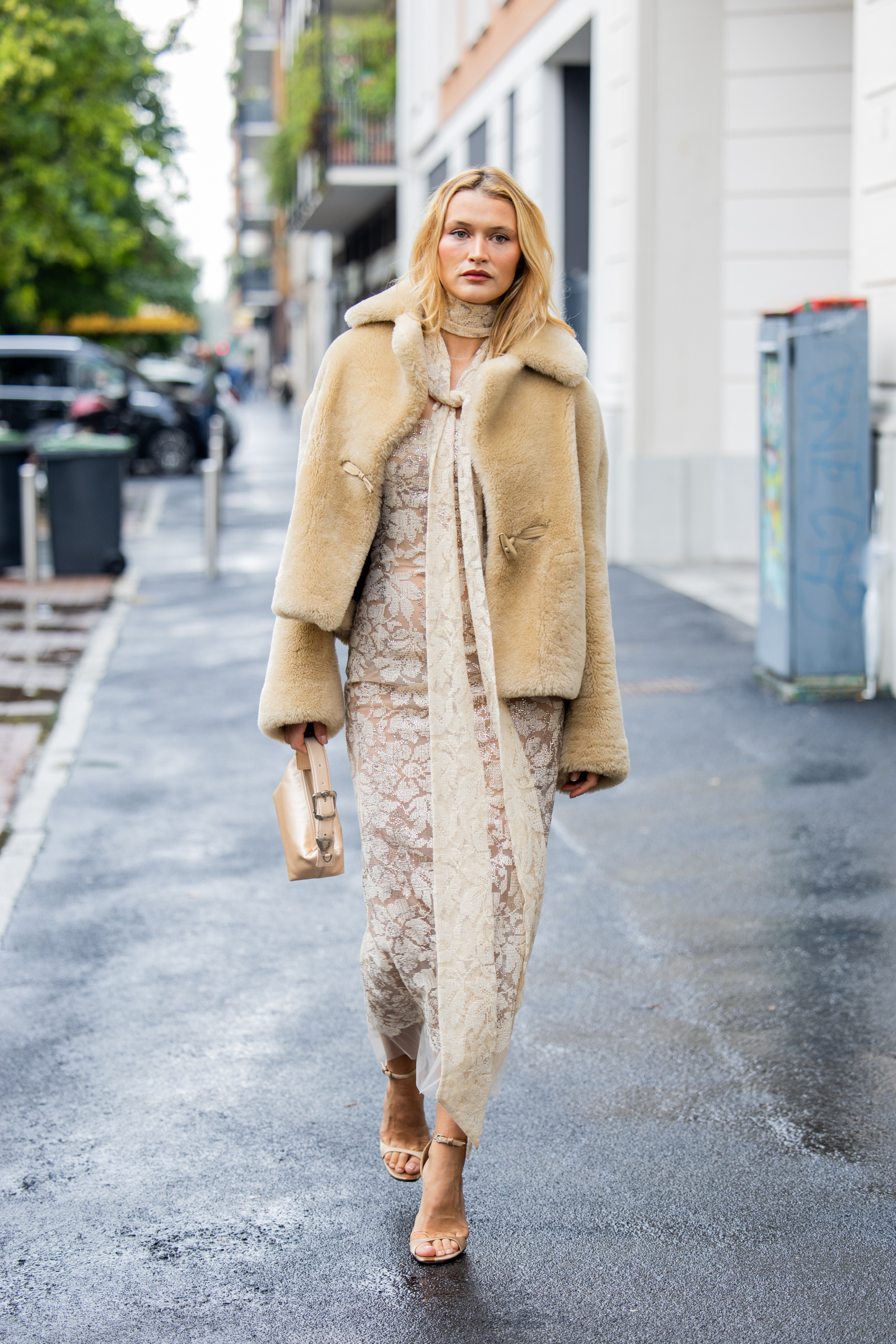 Kobieta spacerująca po ulicy w eleganckiej sukni, otulona kurtką teddy coat w jasnym kolorze.