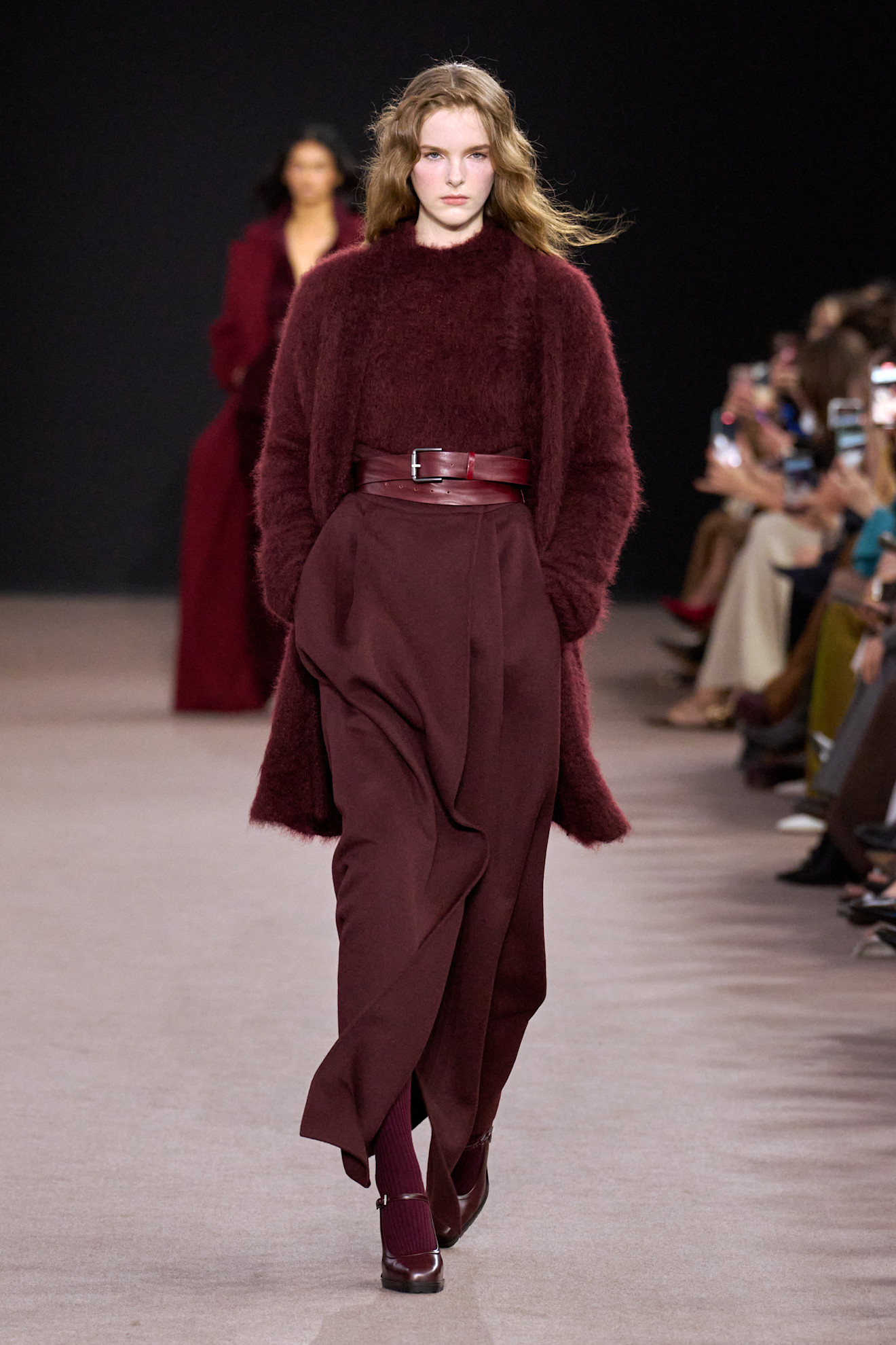 Modelka w obszernym kardiganie w odcieniu burgundowym, nosząca eleganckie spodnie i wysokie buty, na wybiegu.