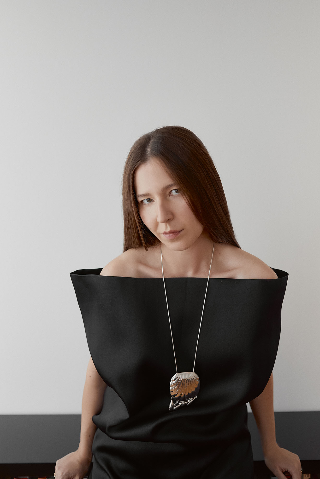 Olivia Kijo w czarnej sukience bez ramion, z ozdobnym naszyjnikiem, siedzi przy minimalistycznym stole.