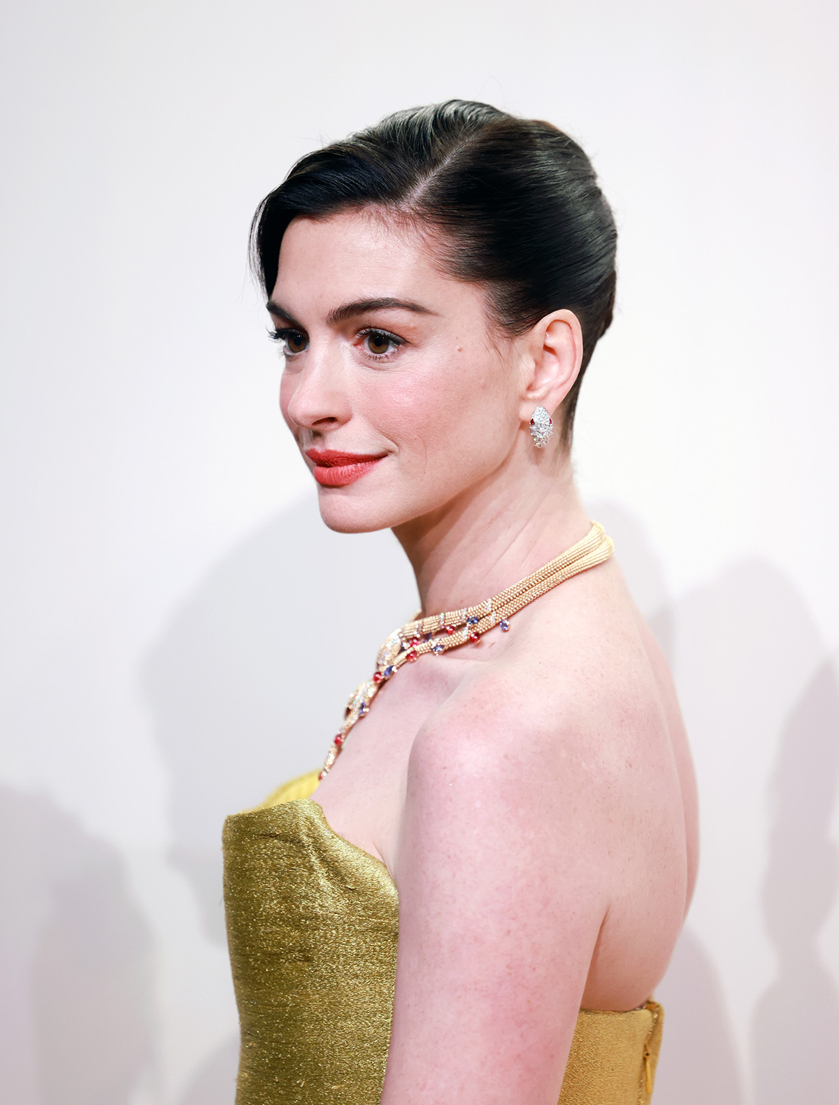 Anne Hathaway z włosami upiętymi w kok i w sukience w kolorze ciemnego złota.