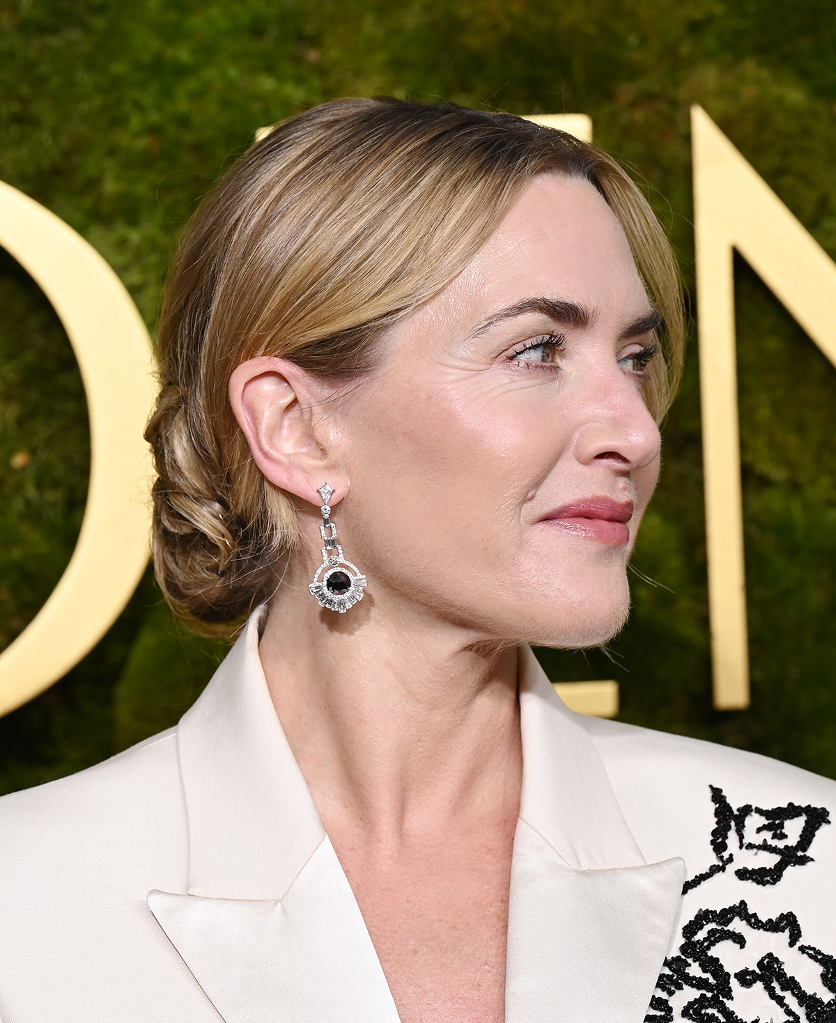Kate Winslet z włosami spiętymi w luźny kok i w białej marynarce z czarnymi ozdobami z koralików.
