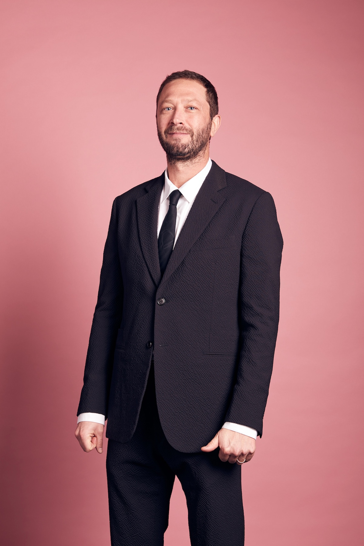 Ebon Moss-Bachrach w eleganckim czarnym garniturze na tle różowego tła, uśmiechając się do kamery.